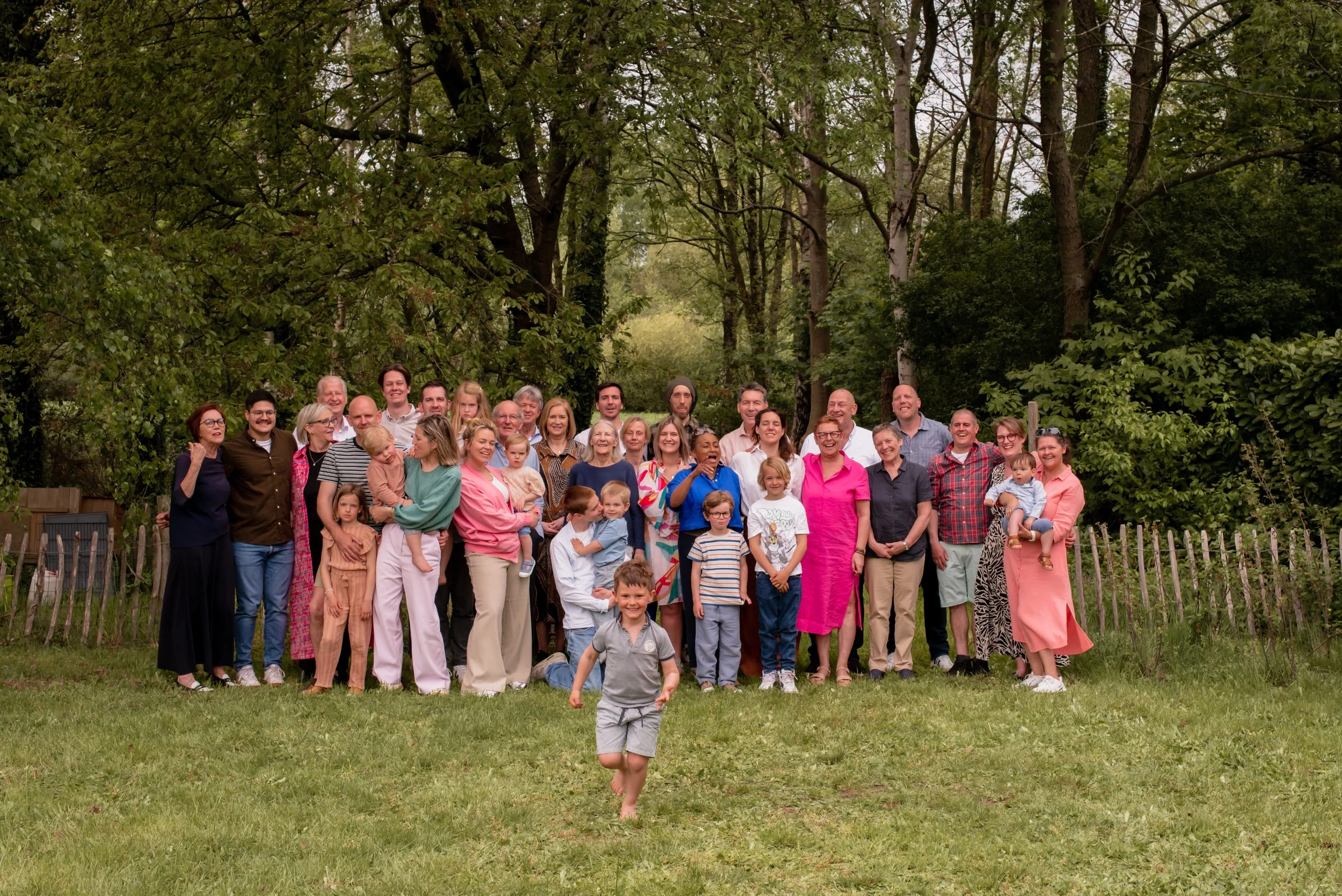 A large group of people, including children and adults, gathered outdoors in a wooded area, with one child running in the foreground.