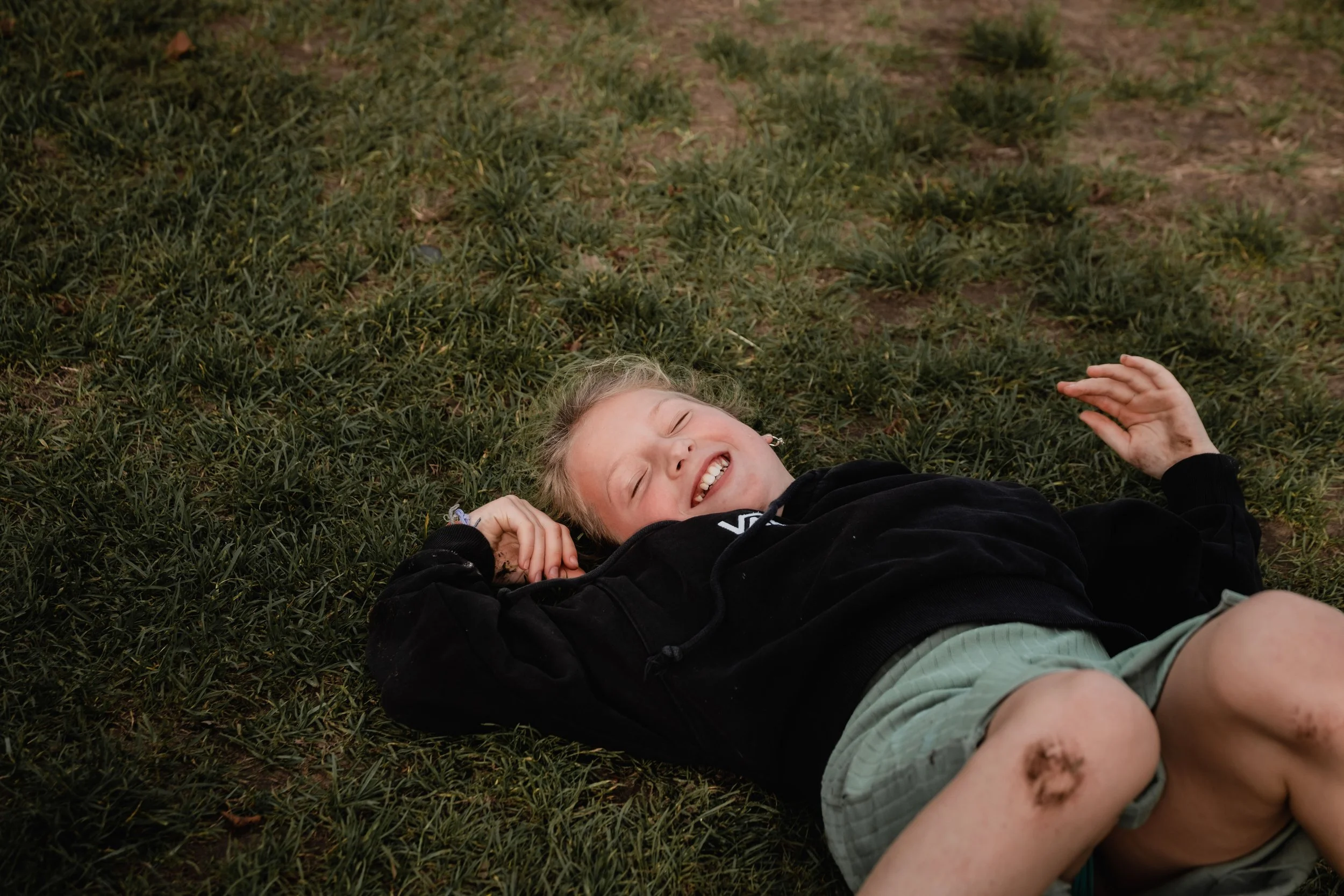 A young child with blonde hair lying on grass and smiling, wearing a black hoodie and gray shorts.