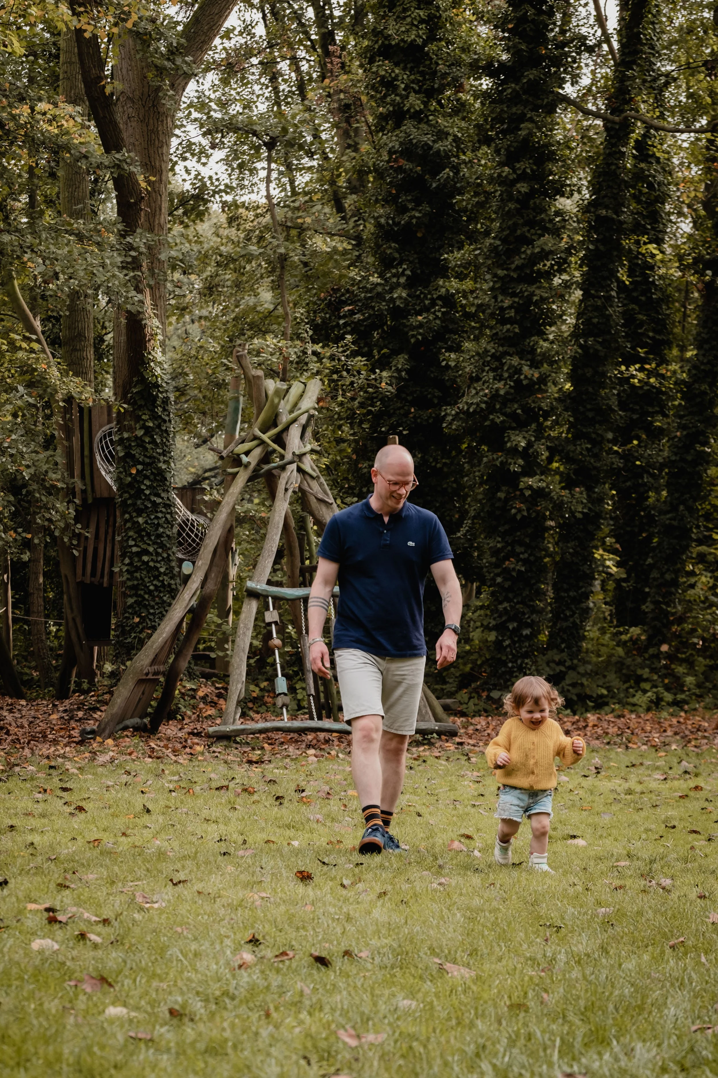 A man and a young girl walking hand in hand in a backyard with a wooden play structure and trees in the background.