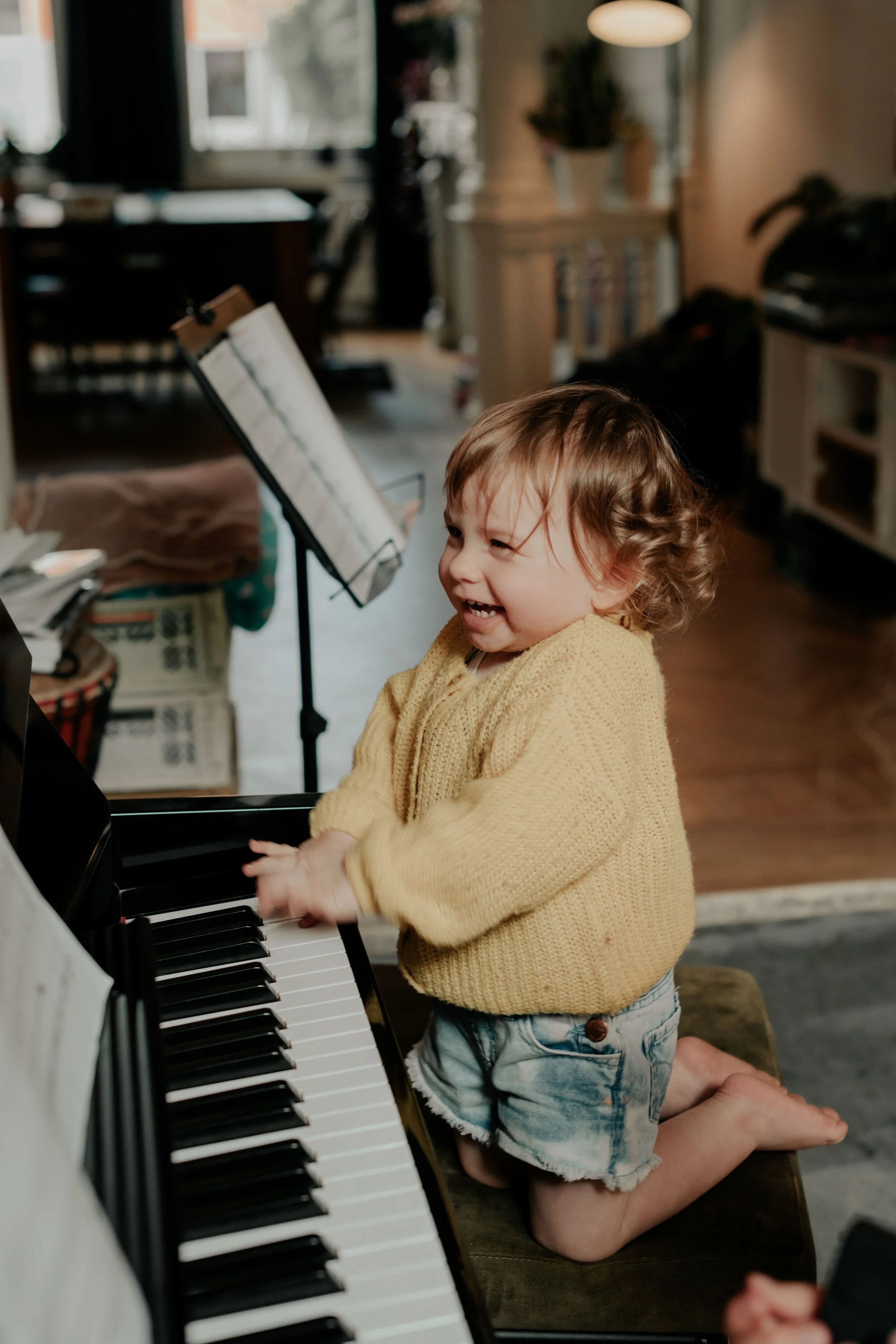 A young child with curly hair wearing a yellow sweater and denim shorts, kneeling on the floor and playing a black grand piano, smiling and enjoying the moment in a cozy, well-lit room.