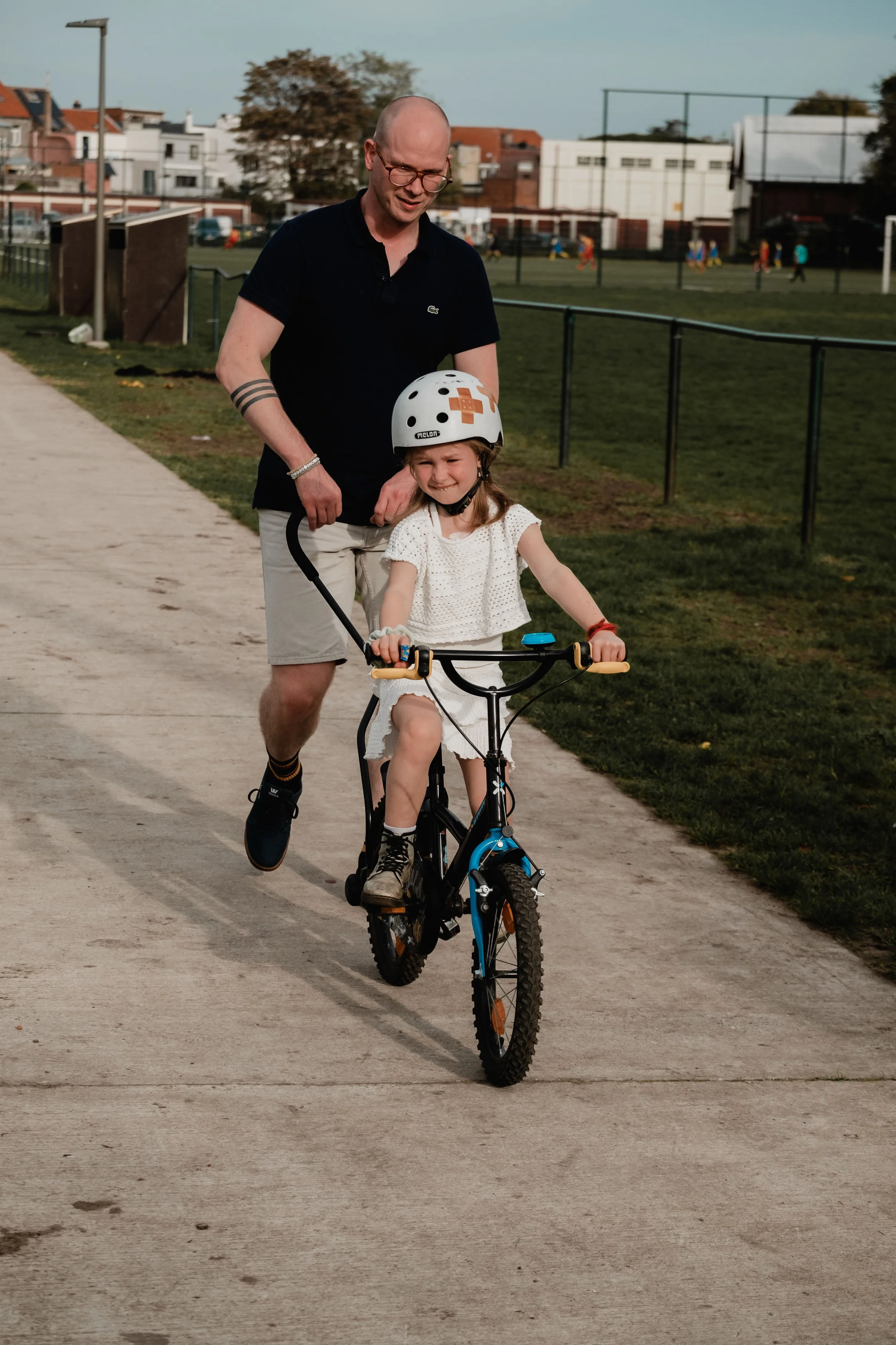 A man and a young girl riding a bicycle together outdoors on a paved path in a park. The girl is wearing a white helmet, a white dress, and is smiling while pedaling. The man, dressed in a black polo shirt and beige shorts, is smiling and guiding her