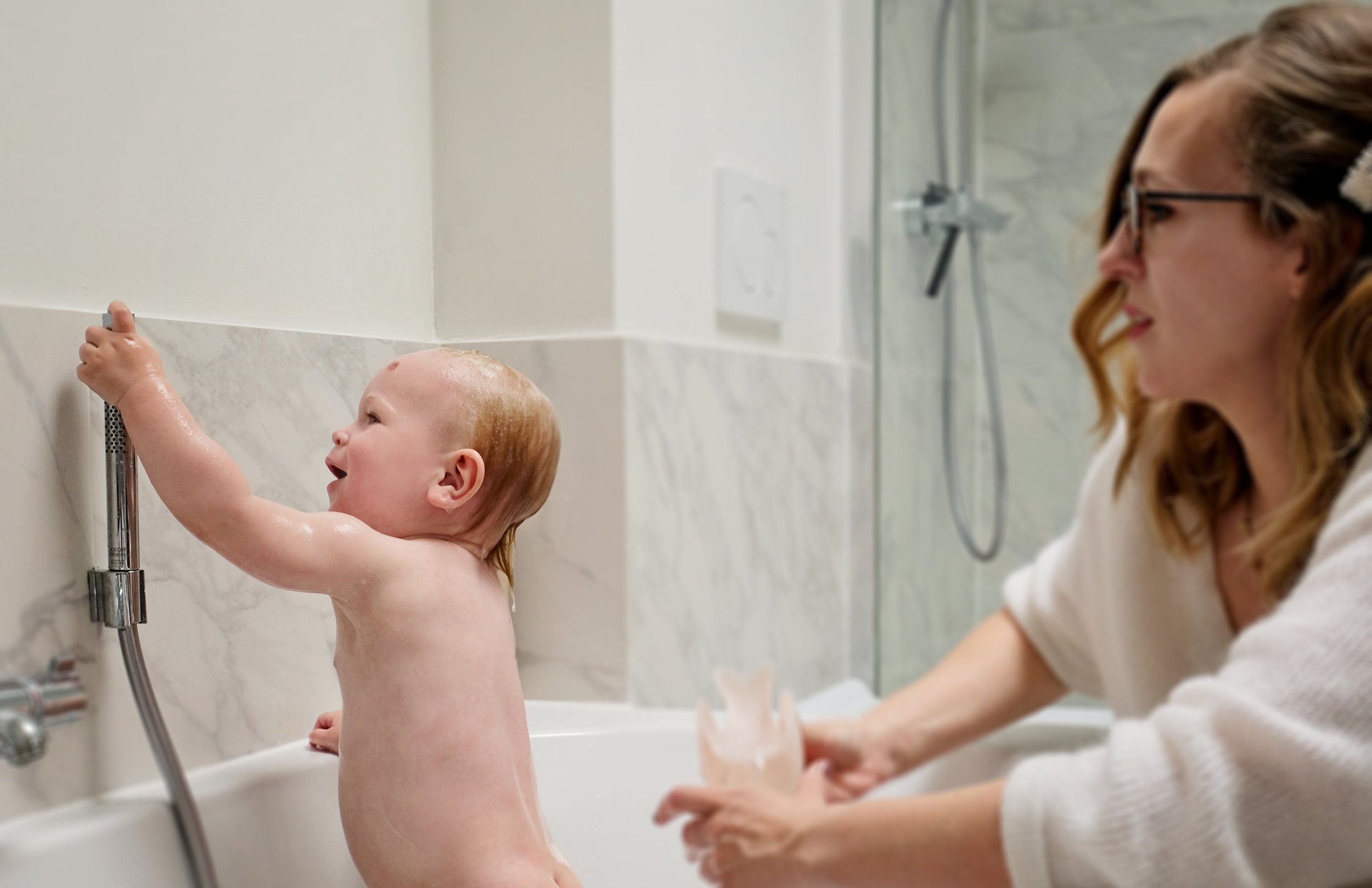 A young child taking a bath in a white bathtub with a woman, likely the mother, smiling and watching. The child has wet hair and is reaching for the showerhead.