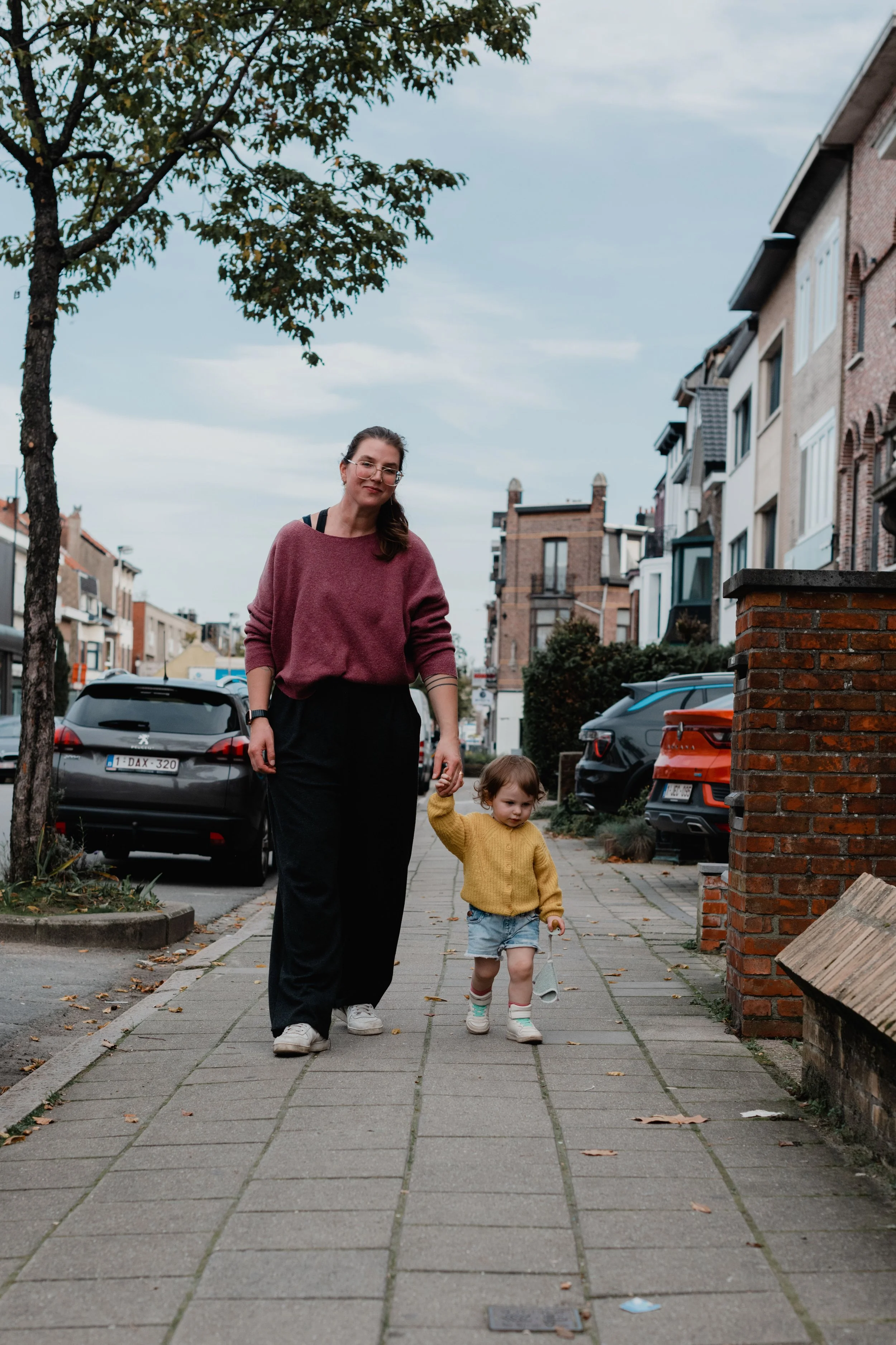A woman and a young girl walking on a city sidewalk with parked cars and residential buildings in the background, under an overcast sky.