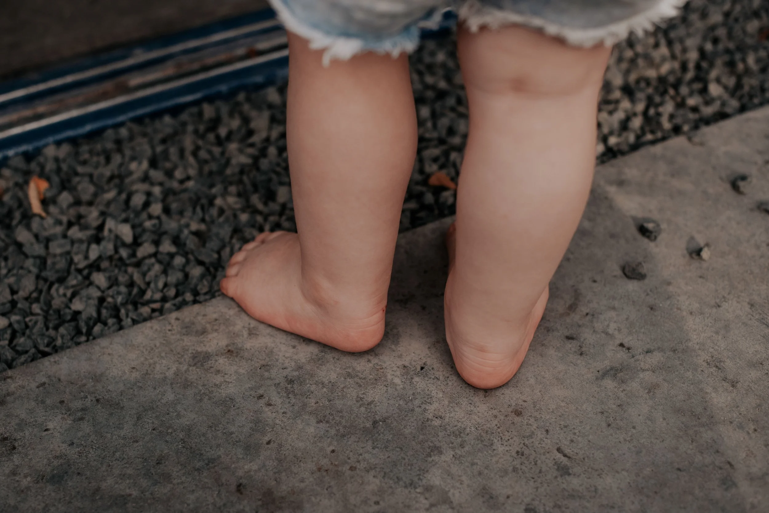 Close-up of a child standing barefoot on concrete next to a gravel surface, wearing frayed denim shorts.