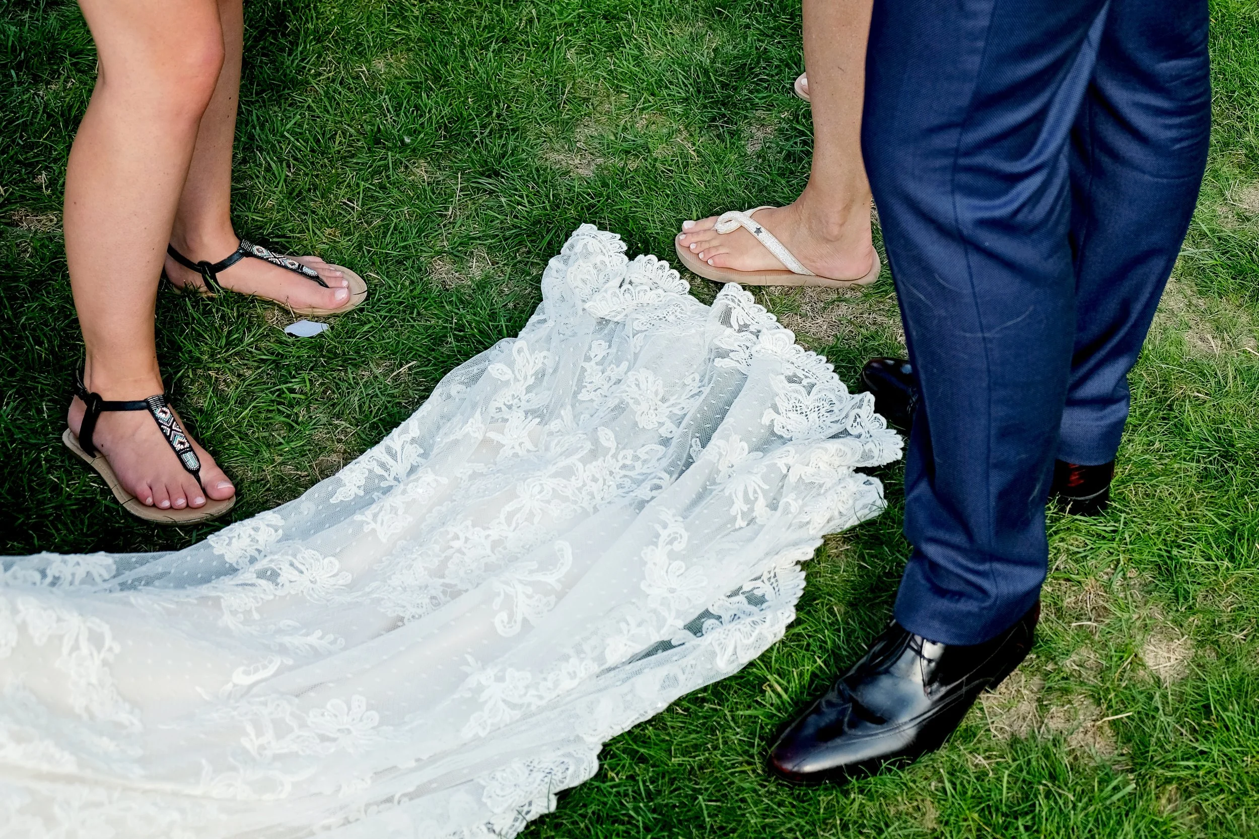 A bride in a white lace wedding dress lying on the grass, with two people standing beside her. The woman is wearing black sandals, and the man is wearing black formal shoes and blue pants. The setting appears to be outdoors on a grassy lawn.