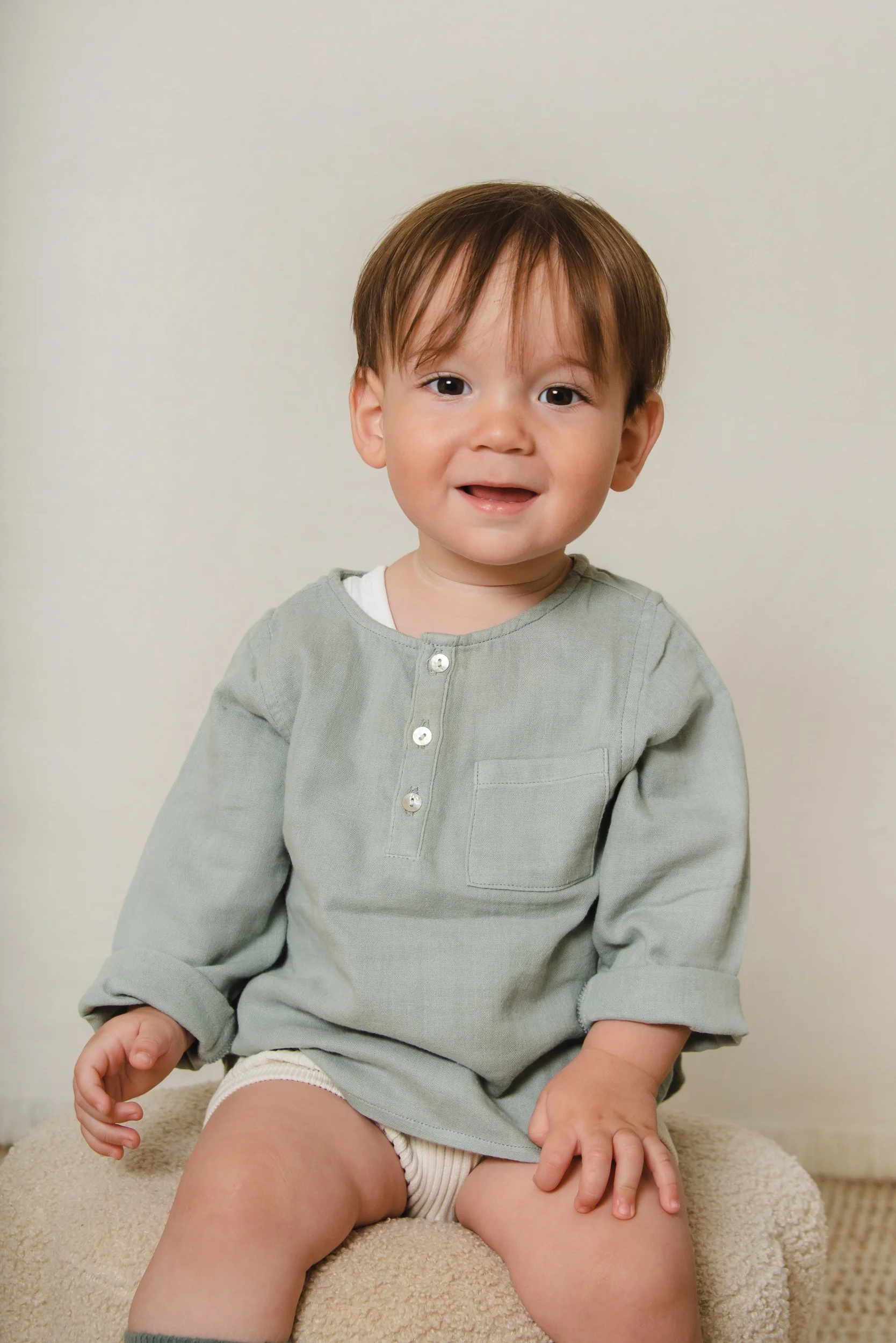A young boy with brown hair and big eyes, wearing a long-sleeved gray shirt and shorts, sitting on a beige cushion against a plain white wall, smiling softly at the camera.
