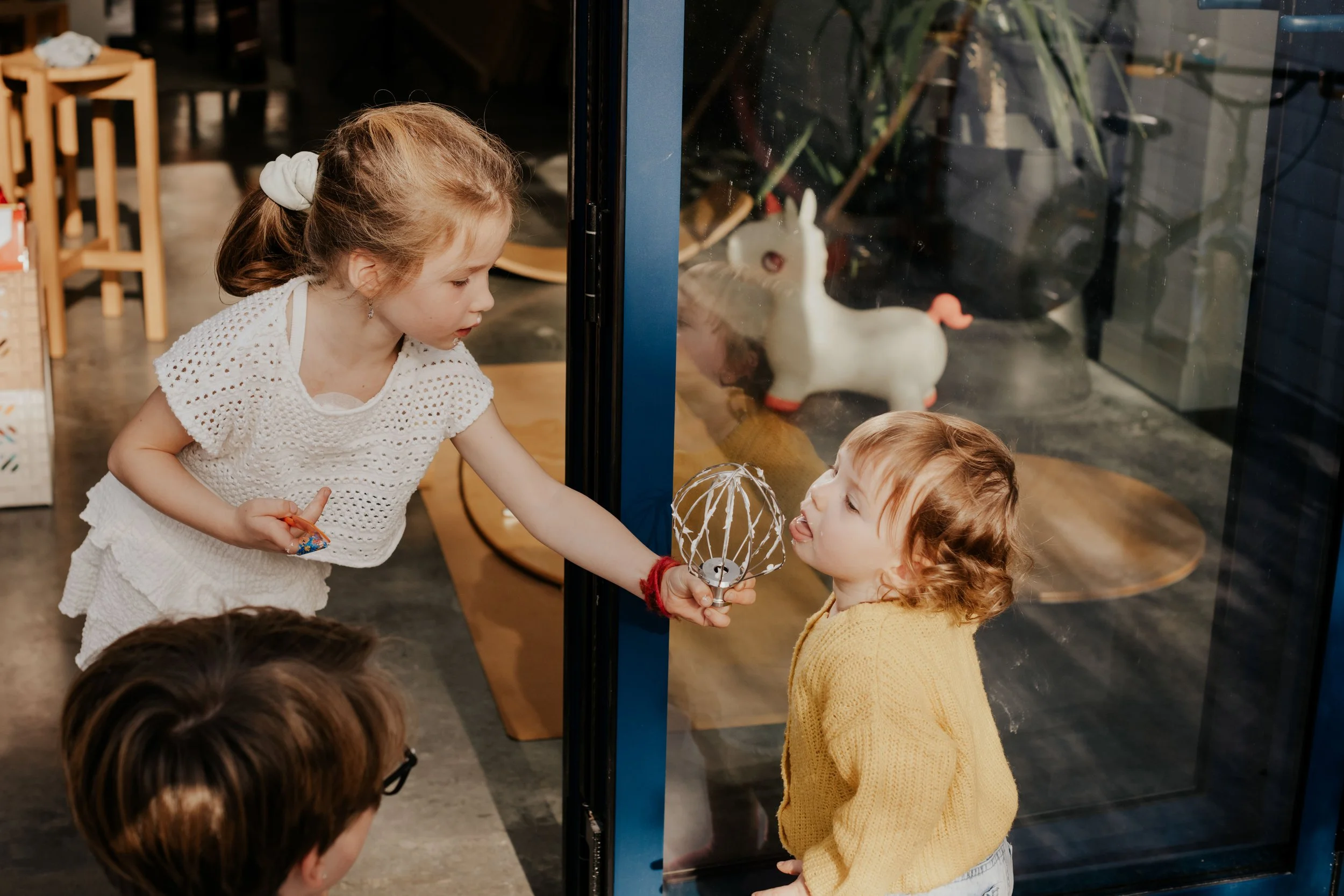 A young girl with curly red hair and a yellow sweater observes a white ceramic unicorn figurine through a glass window, while another girl with blonde hair and a white crocheted top leans in to look, with other children partially visible in the foreg