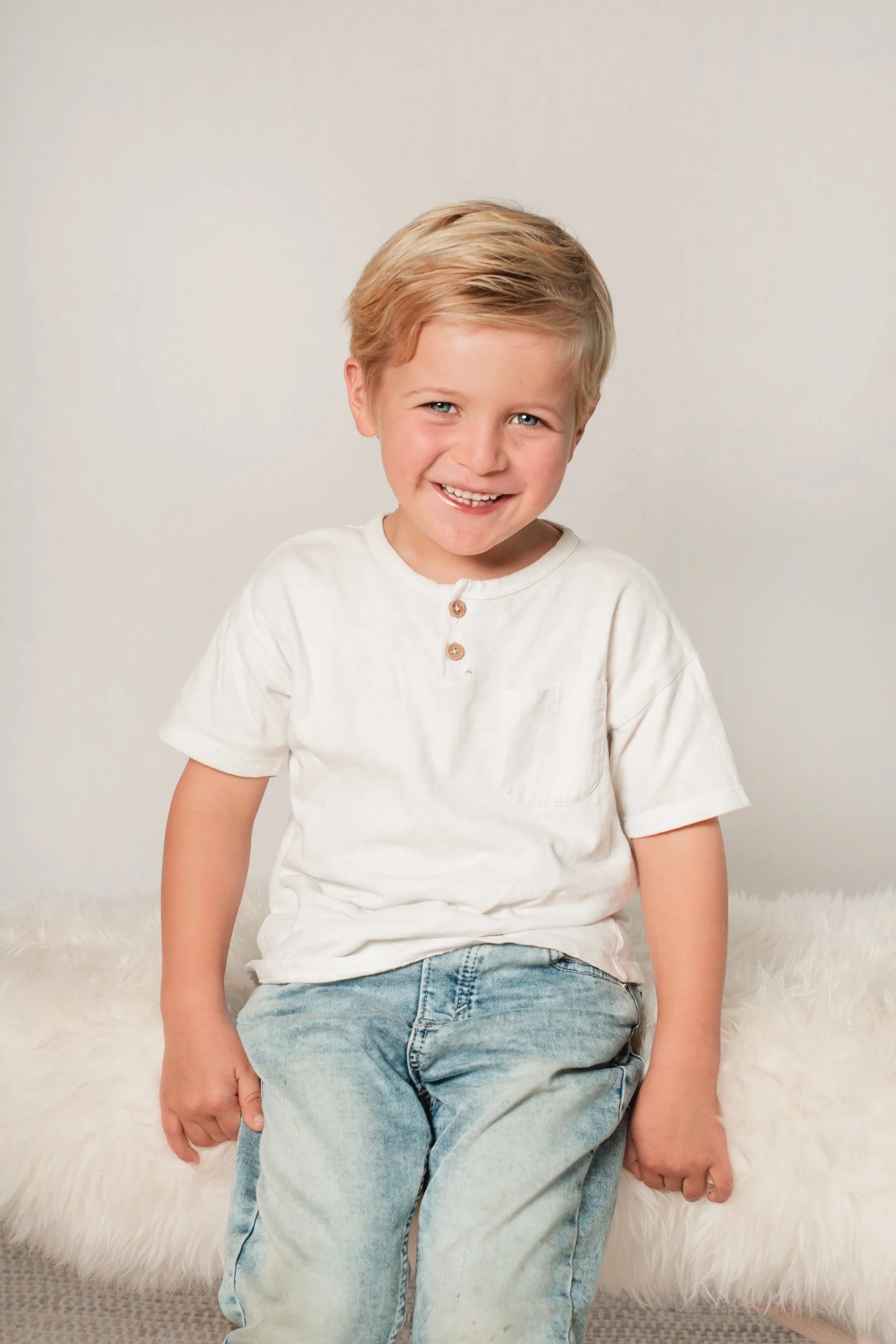 A young boy with blonde hair and blue eyes, smiling, wearing a cream-colored T-shirt with three buttons and light-wash jeans, seated on a fluffy white surface with a plain light-colored background.