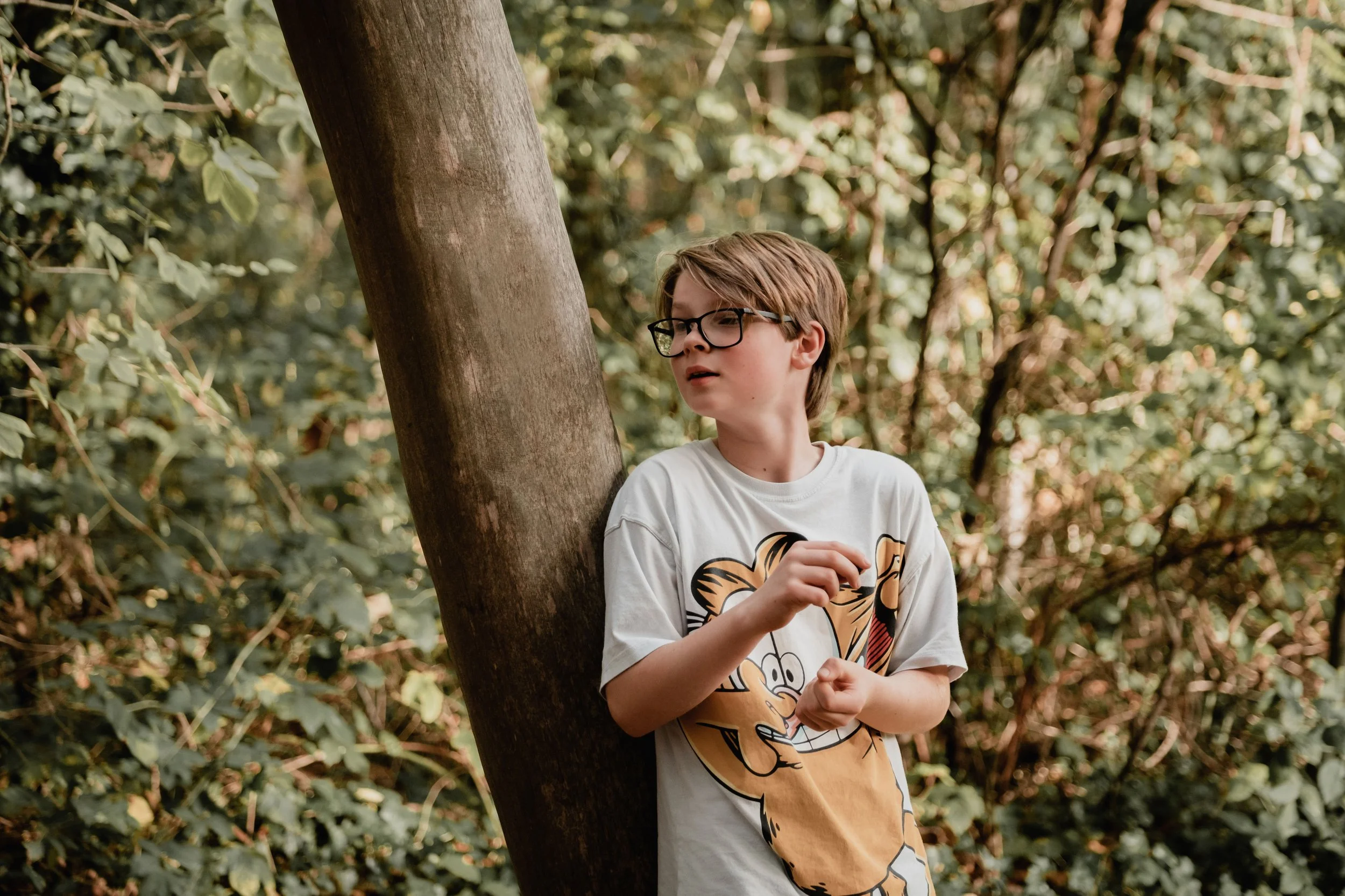 A young boy with glasses and light brown hair, wearing a white T-shirt with a cartoon character, stands next to a tree in a forested area with green foliage.
