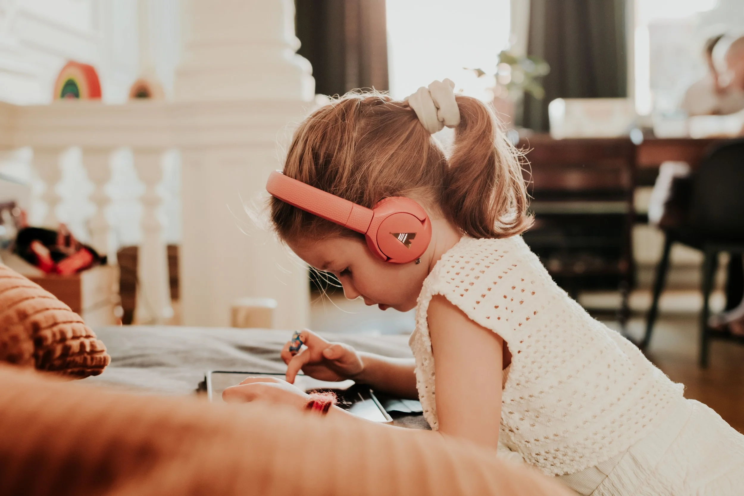 A young girl with red hair and a cream-colored hair bow wearing pink headphones, sitting at a table and playing with a tablet.