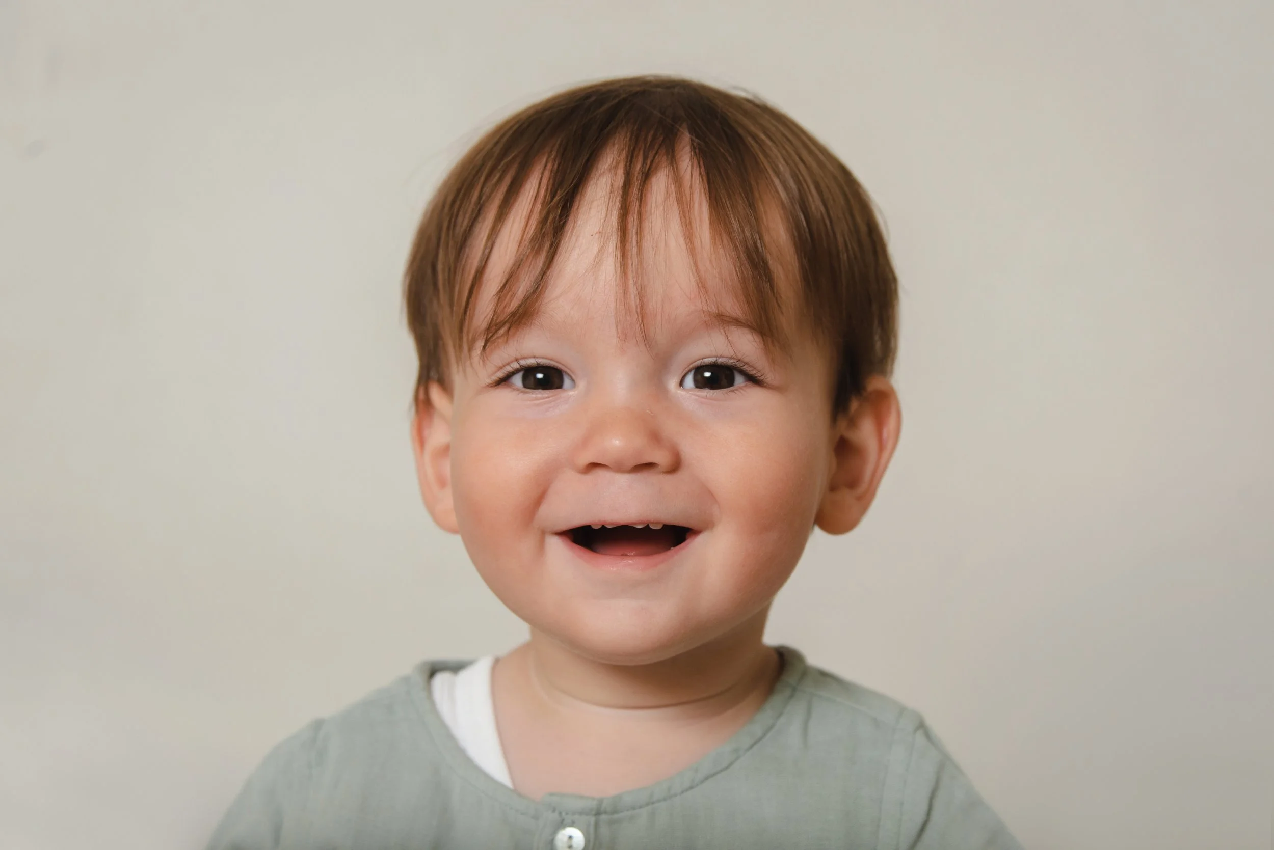 A young boy with brown hair and brown eyes smiling with his mouth open, wearing a gray shirt with a white collar, against a plain light-colored background.