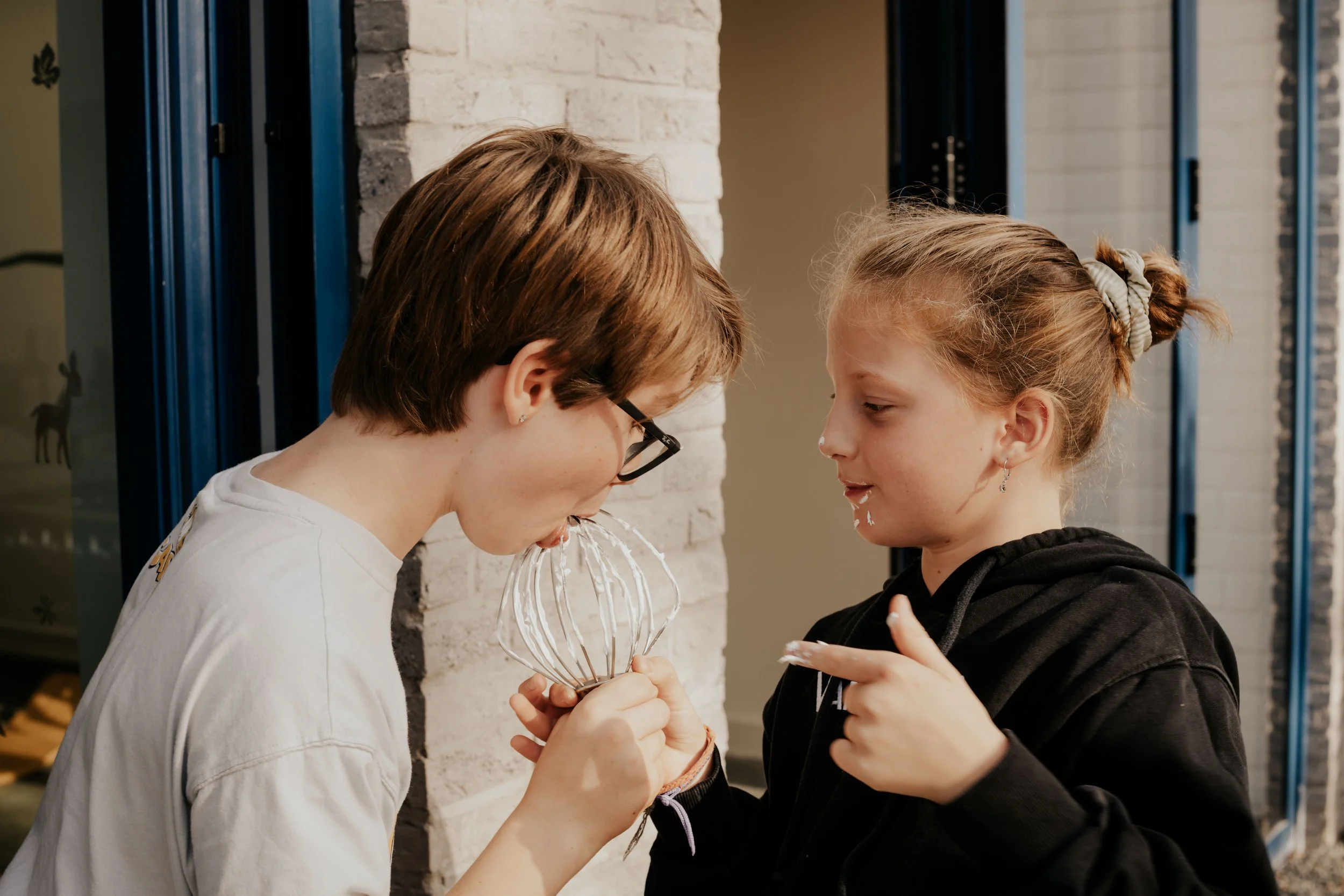 Two young girls are touching foreheads and holding a white whisk together near a brick wall and a window.