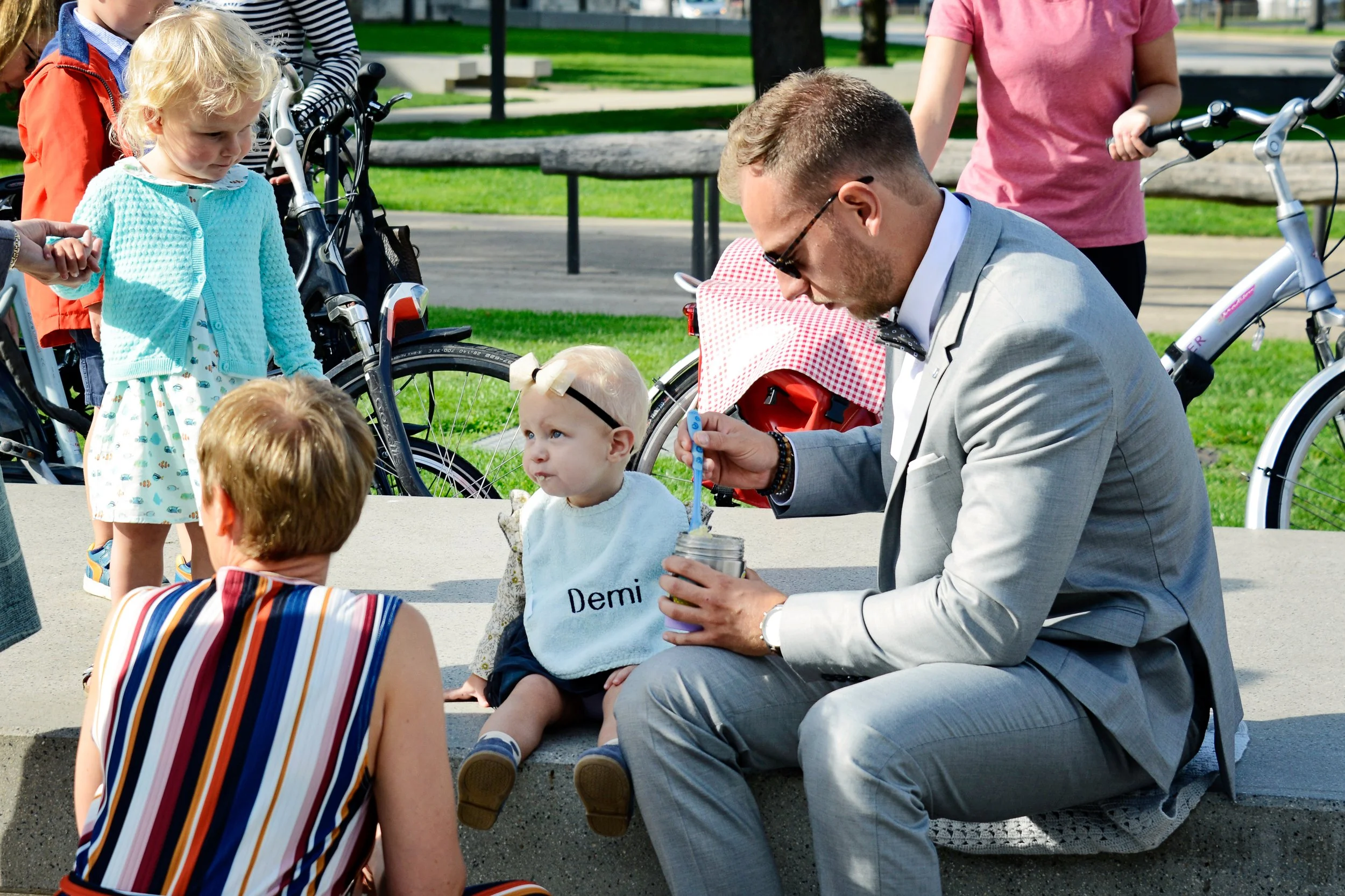 A man in a gray suit and sunglasses sits on the ground, holding a jar with a spoon. A young girl with a blue sweater and a white dress with colorful patterns stands nearby. Several children and adults are around, some on bicycles, in a park with gree