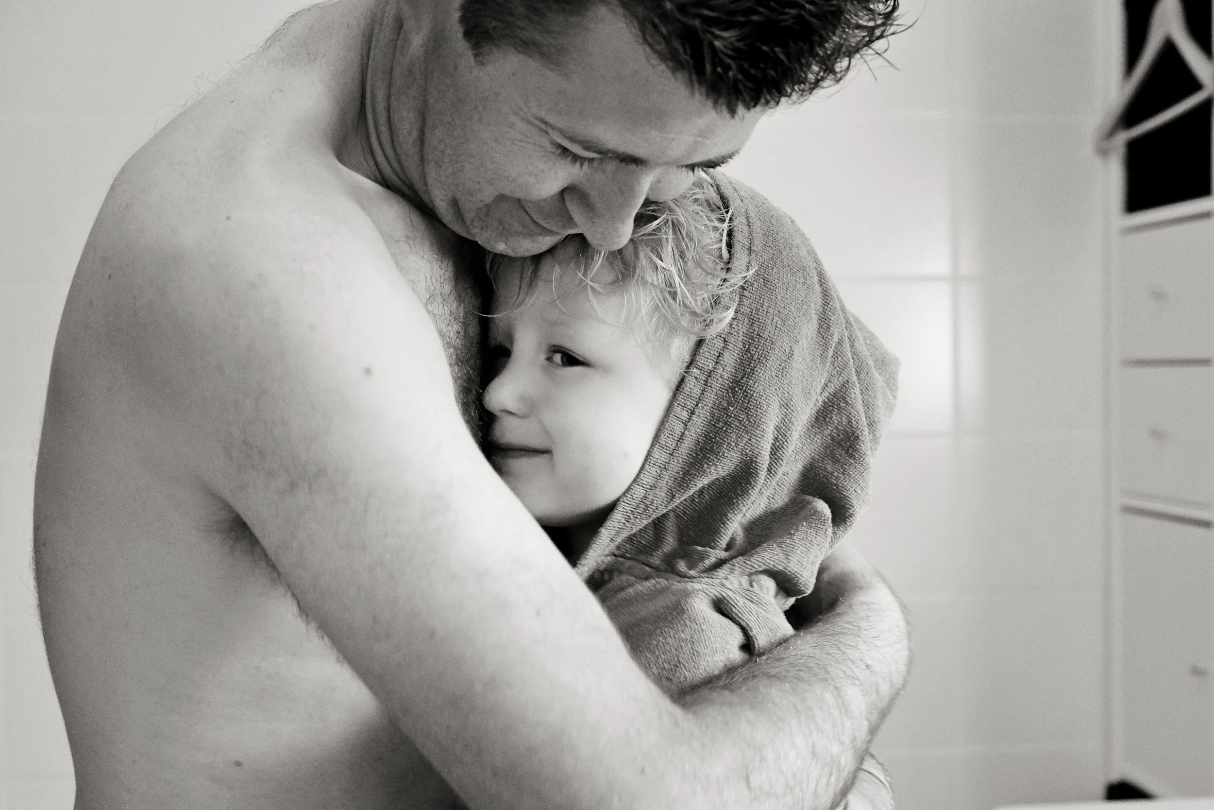 A black and white photo of a man and a young girl hugging closely, both with eyes closed. The man is shirtless and the girl is wrapped in a towel. The scene appears tender and intimate.