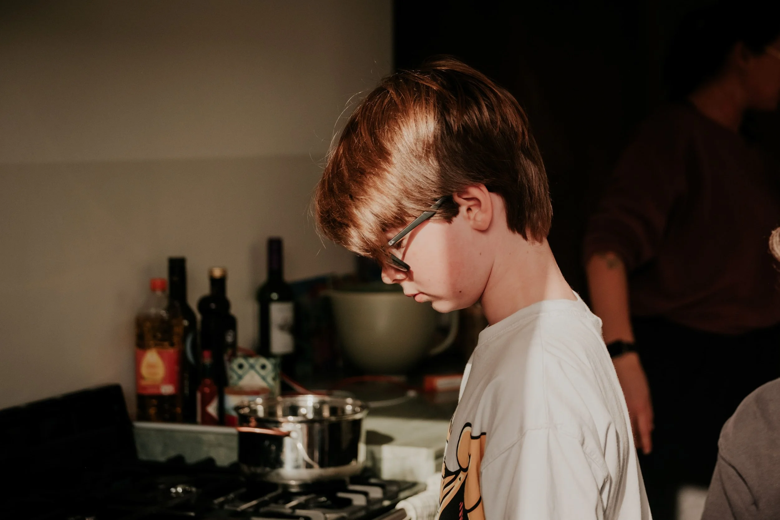 A young boy with brown hair and glasses looking down in a kitchen, with various bottles and kitchen utensils in the background.