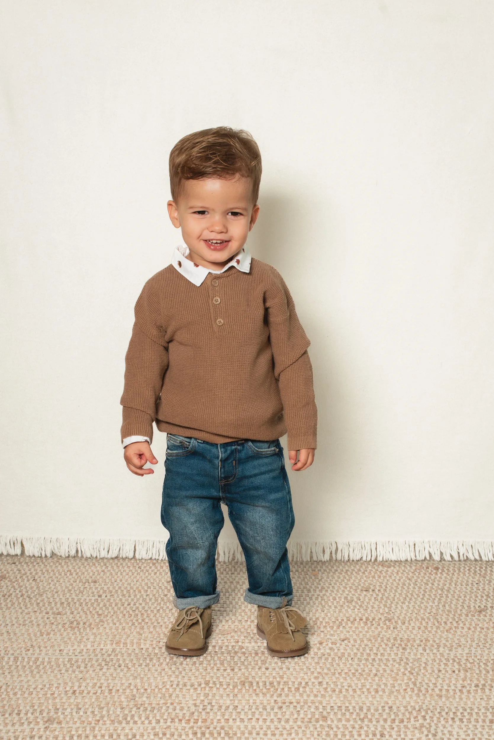 A young boy with light brown hair wearing a light brown sweater, white collared shirt, blue jeans, and tan boots, standing on a beige patterned rug against a plain white wall.