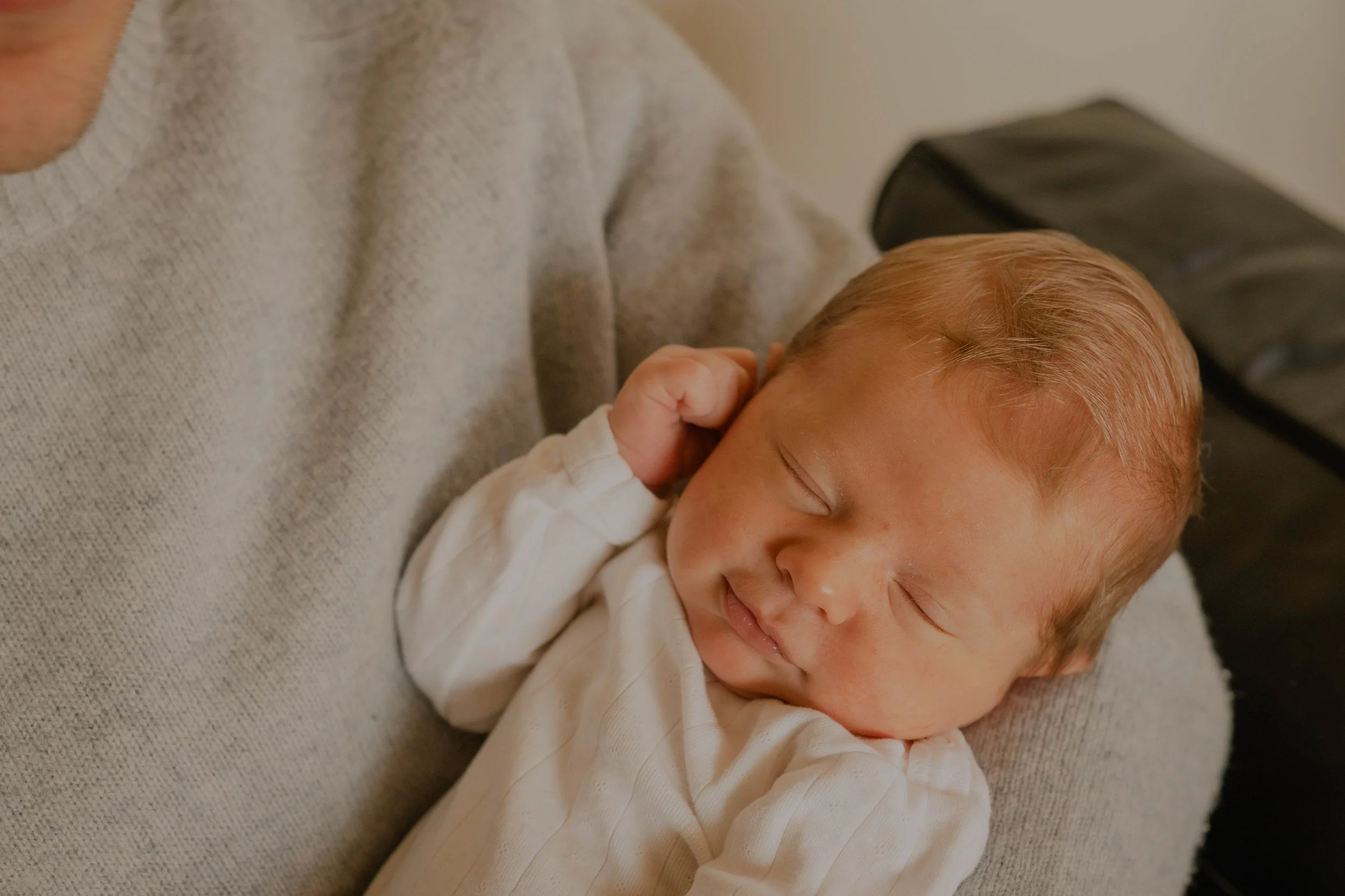 A young child with light skin and reddish hair is peacefully sleeping while resting on an adult's arm, who is wearing a light gray knit sweater.
