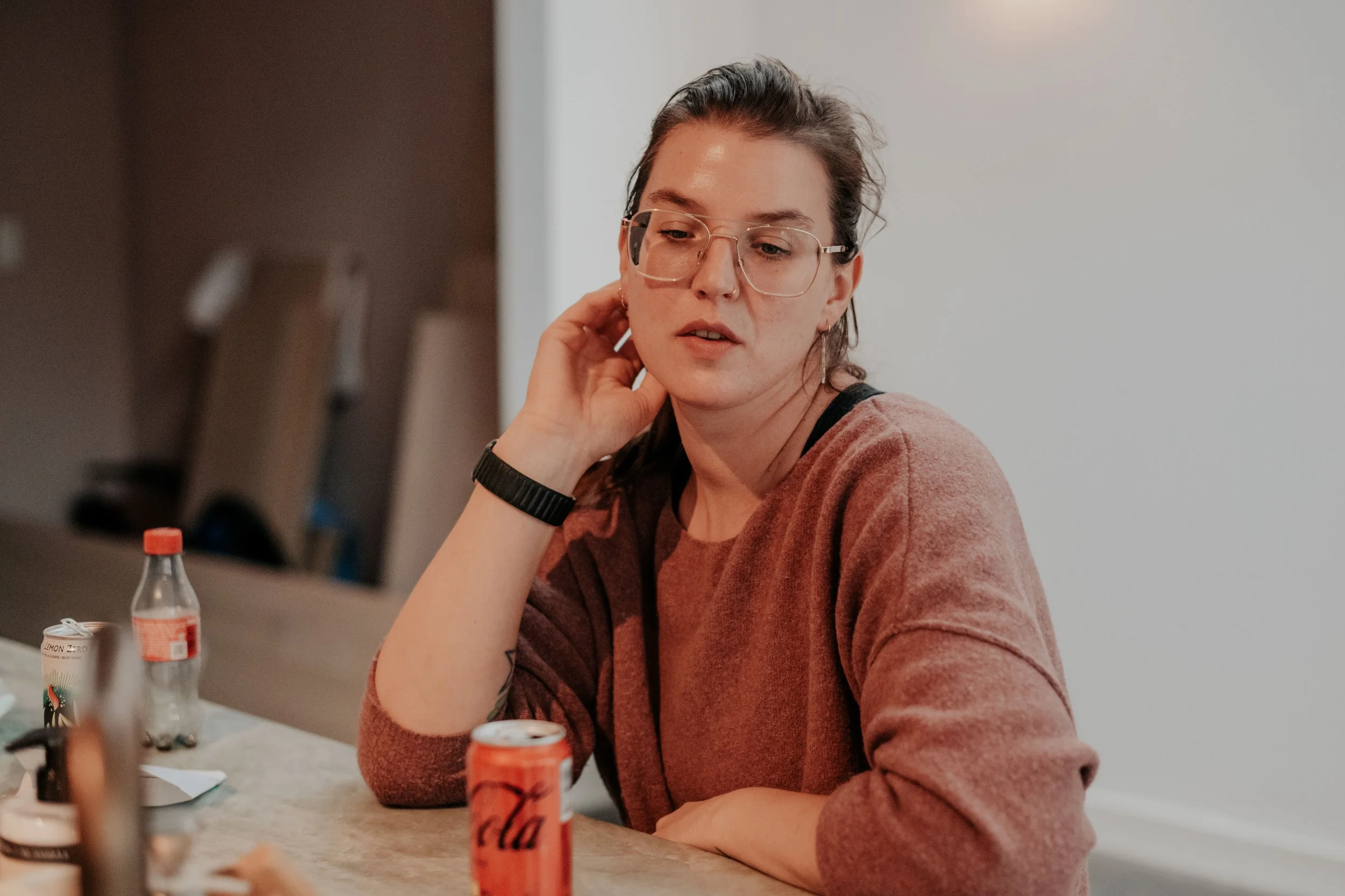 A woman with glasses and a brown sweater sitting at a table, looking at a computer screen, with bottles and cans on the table.