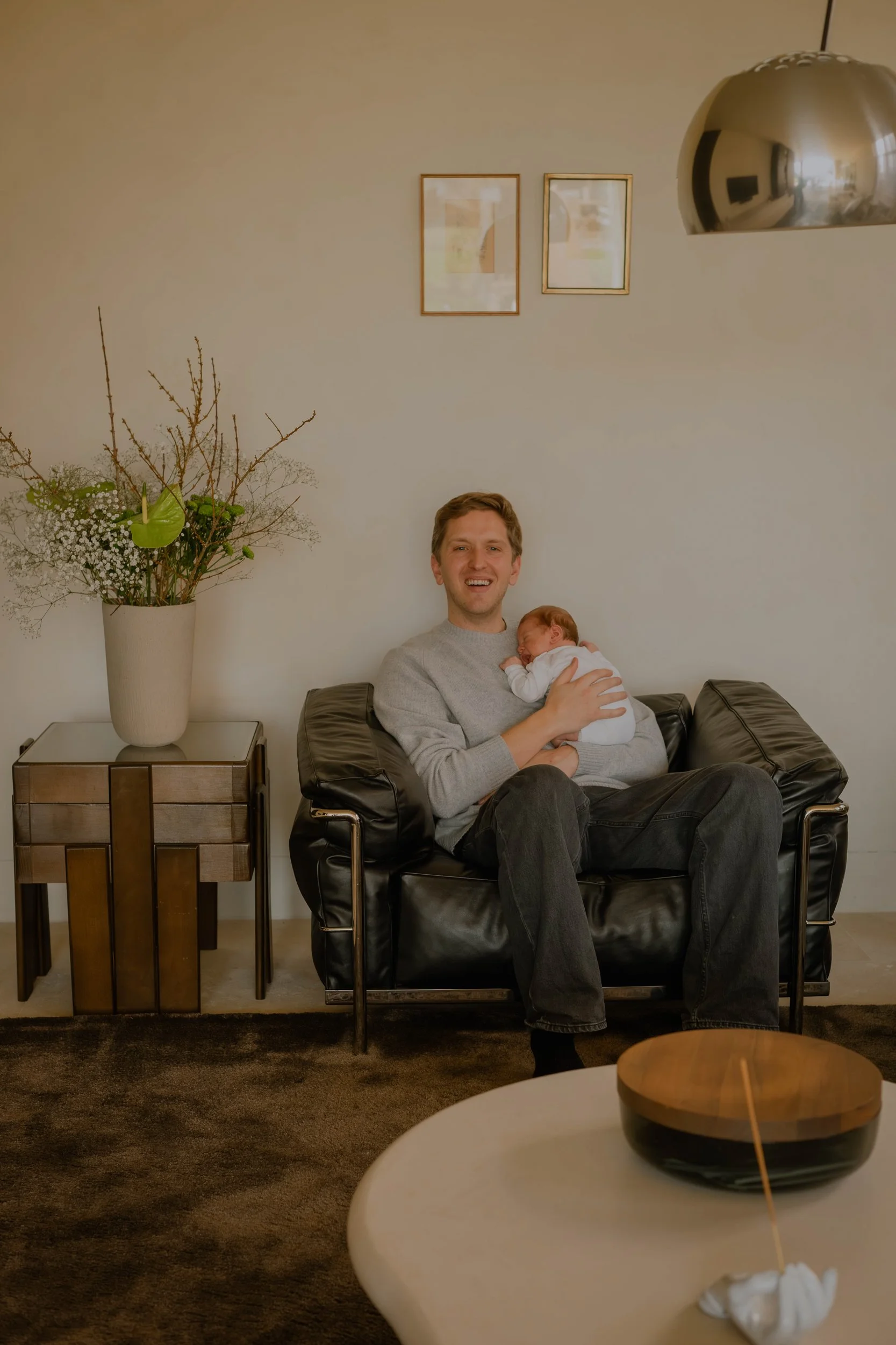 A man sitting on a black leather chair holding a sleeping baby in a white outfit. A large beige flower arrangement on a side table to the left, two framed pictures on the wall behind, and a reflective metallic hanging lamp. In the foreground, a white