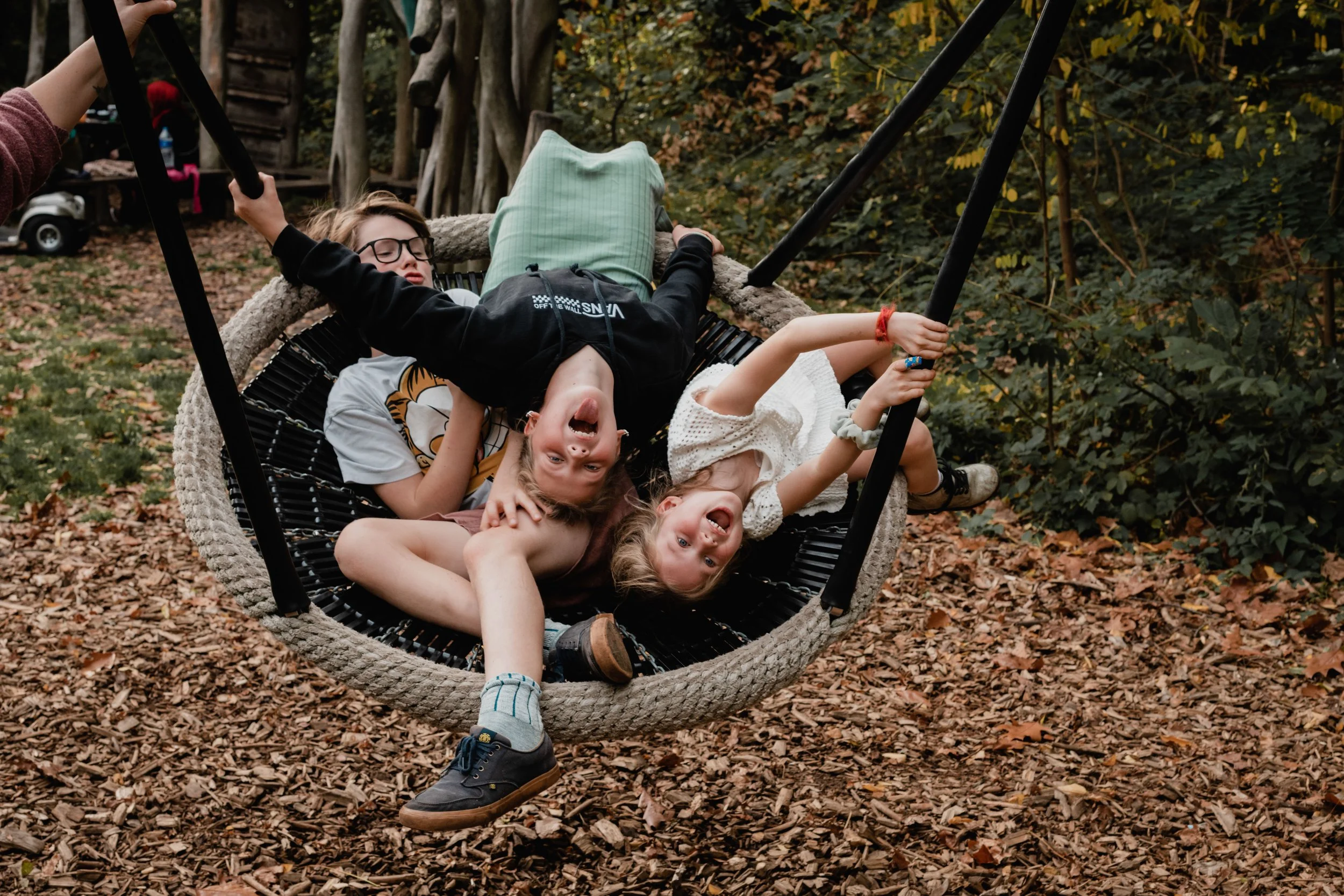 Children playing and laughing on a large outdoor swing in a wooded park during fall.