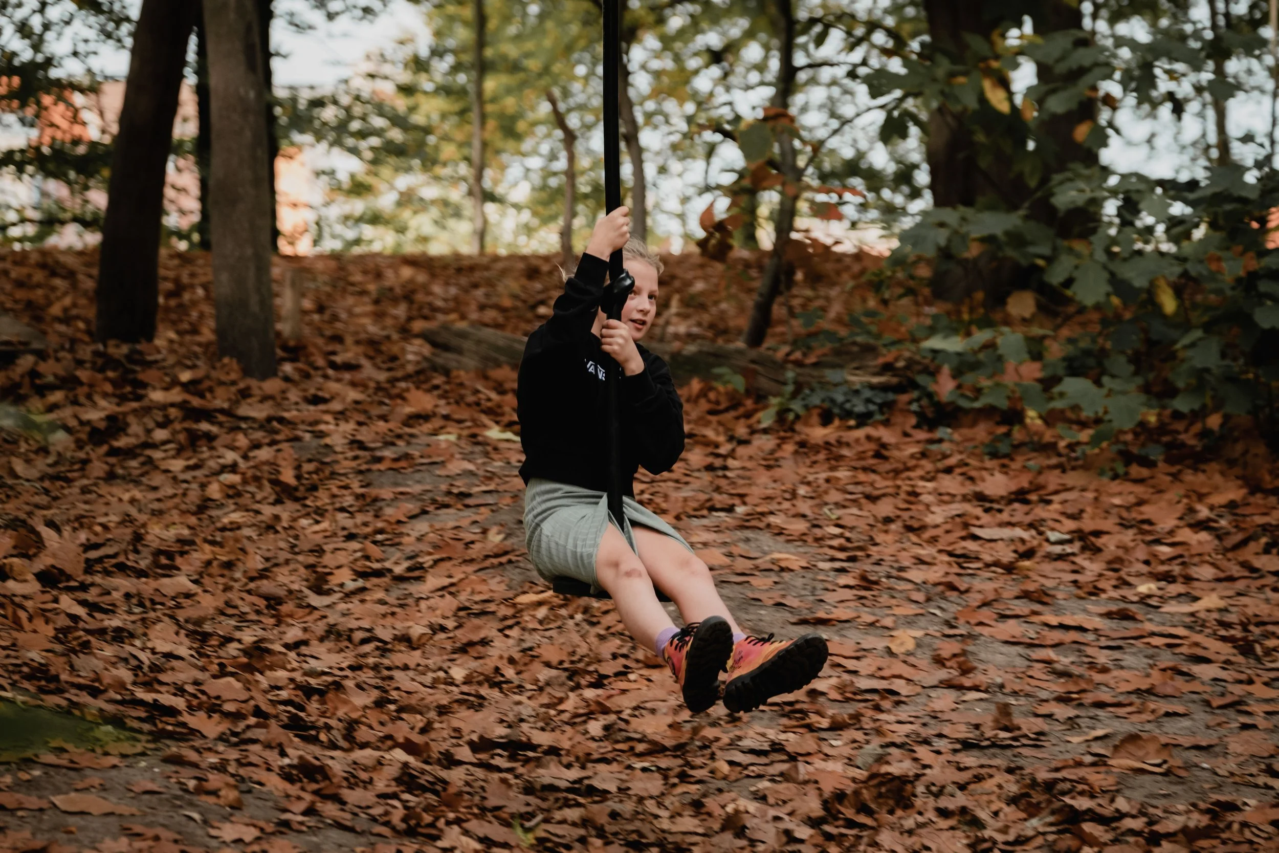 A young girl with blonde hair wearing a black hoodie and gray shorts is sitting on a swing in a wooded area during autumn, with fallen leaves on the ground and trees with green and orange leaves in the background.