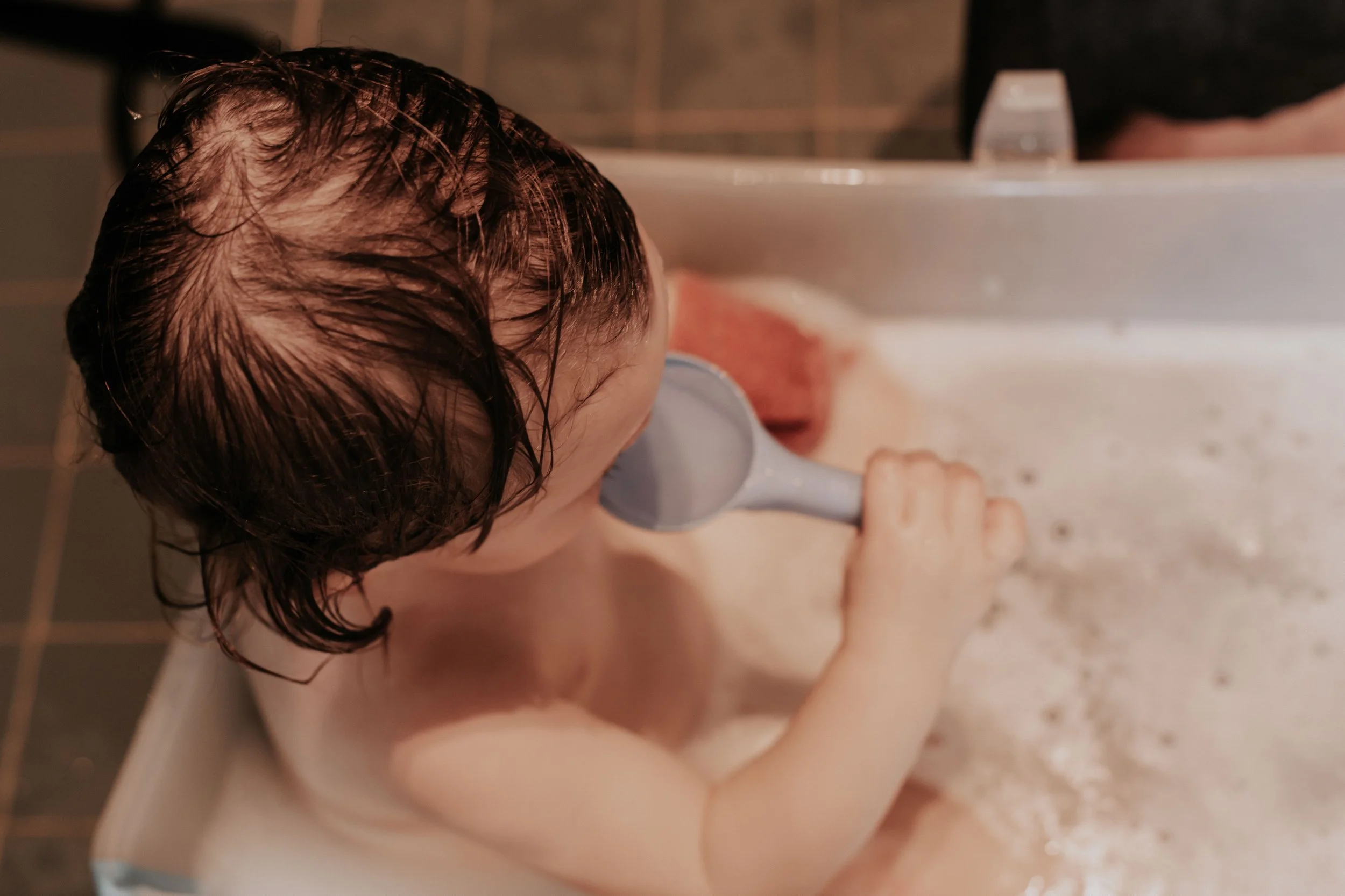 Child taking a bath with a handheld showerhead, wet hair visible, in a tiled bathroom.