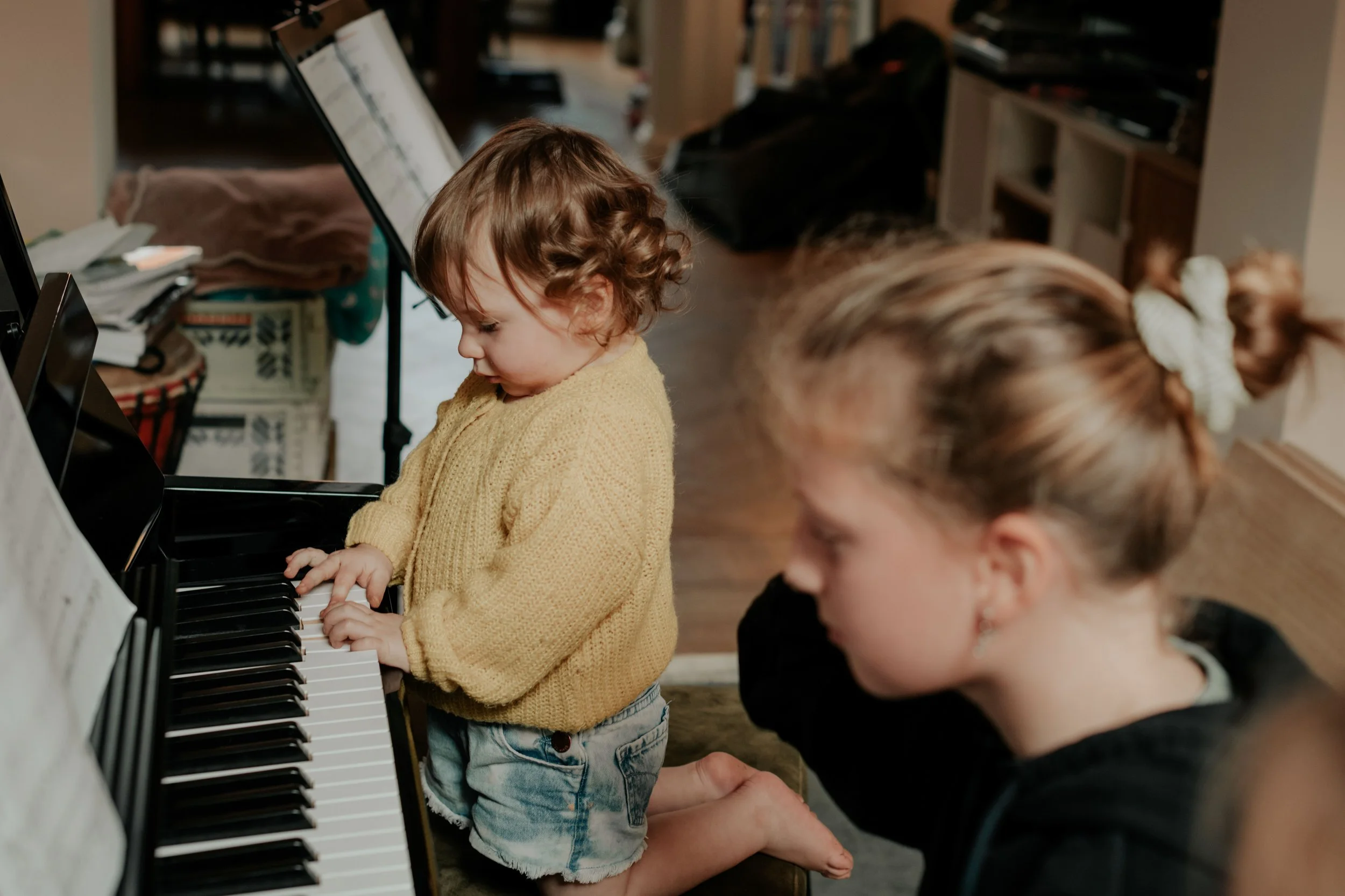 A young girl in a yellow sweater playing the piano with another child watching closely.