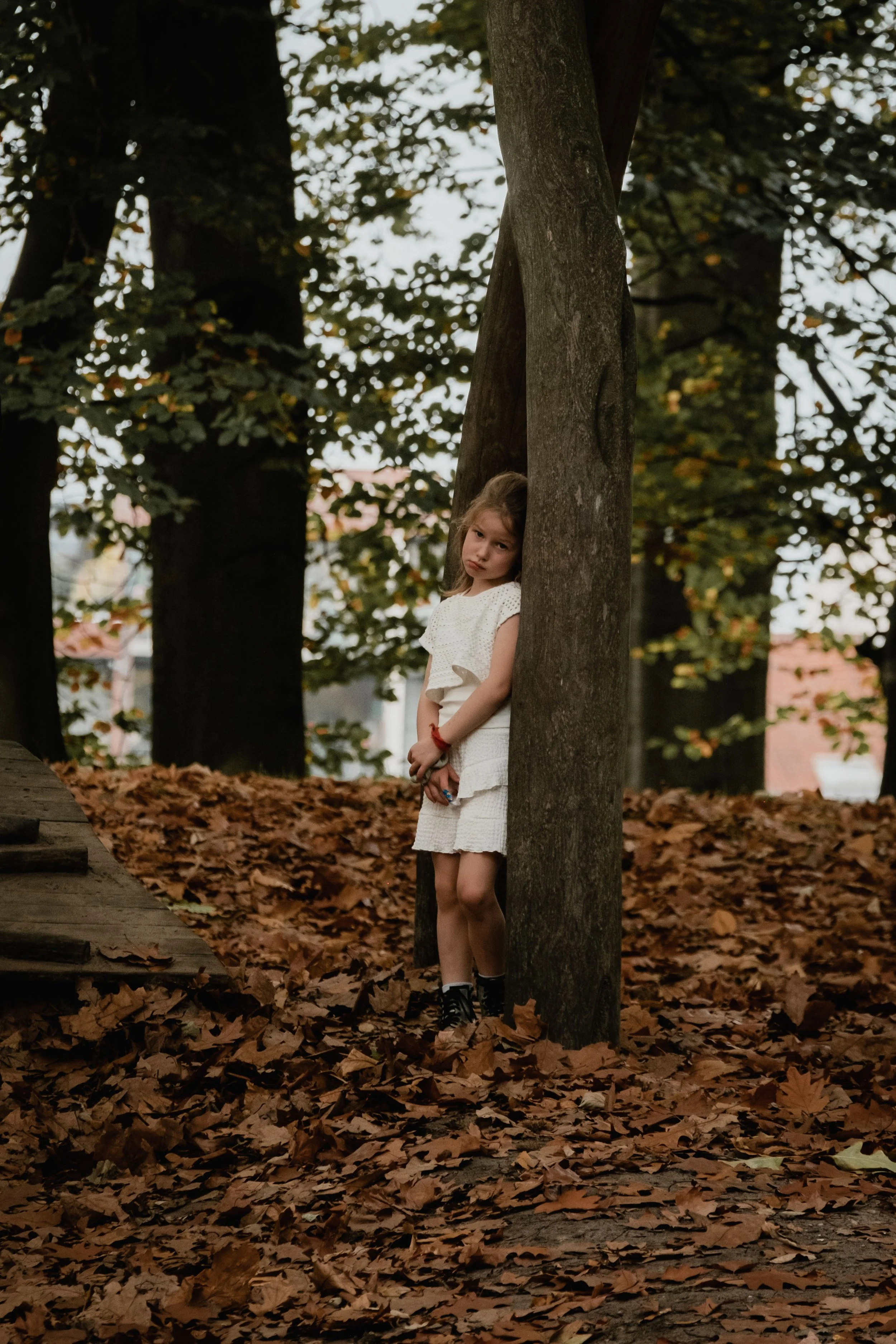 A young girl leaning against a tree in a park with fallen leaves on the ground and trees in the background.