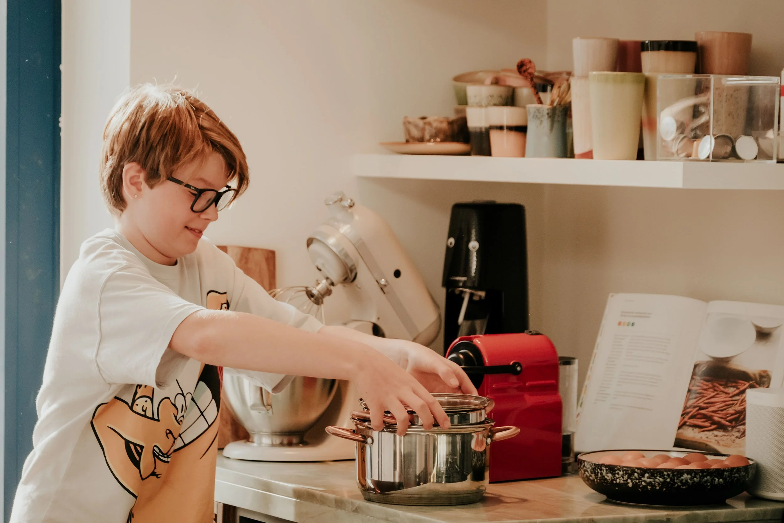 A young person with short reddish hair and glasses is cooking at a kitchen counter. They are wearing a white shirt with a cartoon graphic. They are stirring or working with a metal canister. The kitchen shelves behind them have various mugs, bowls, a