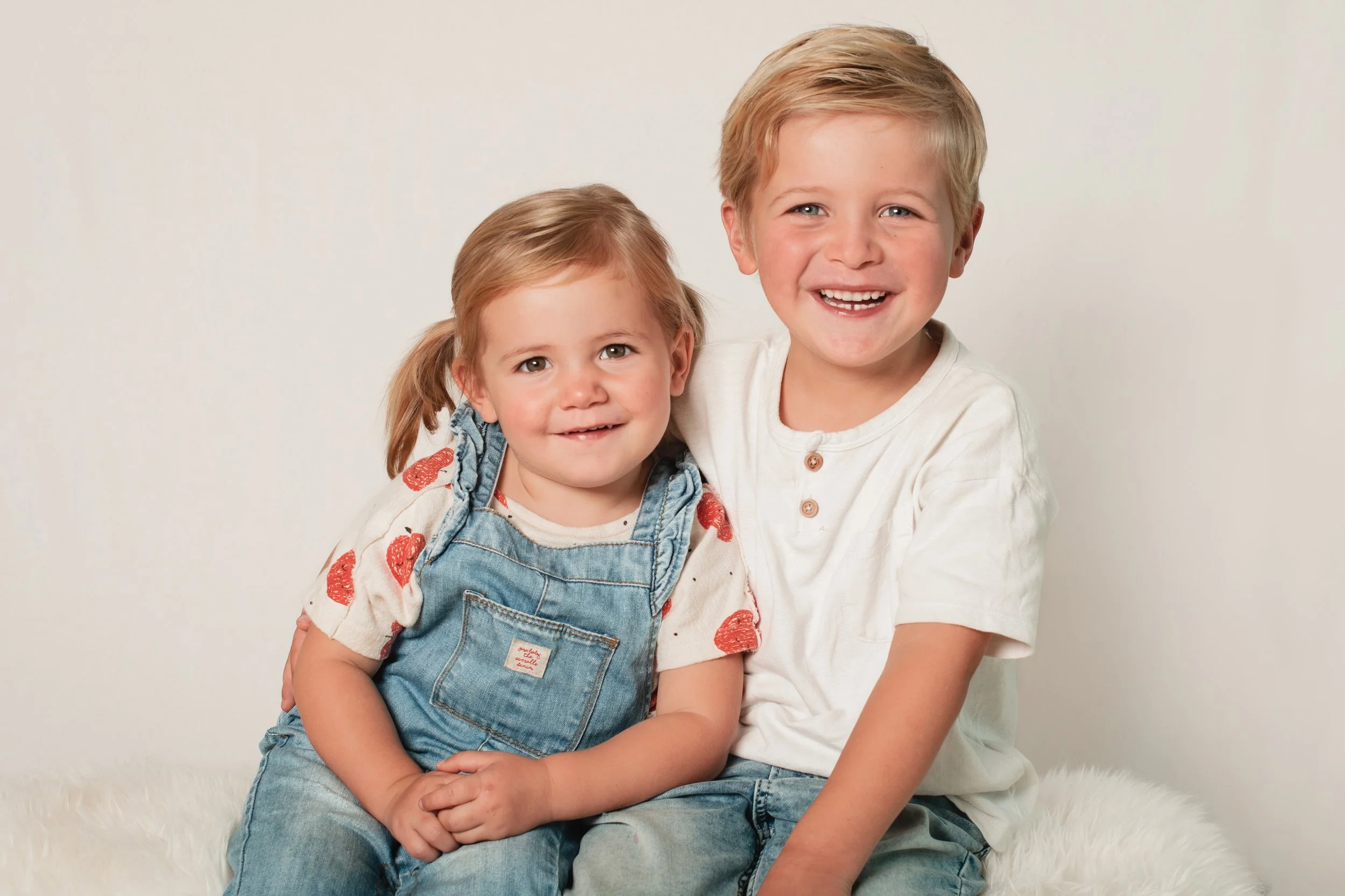 Two young children, a girl and a boy, sitting together on a fluffy white surface against a plain light background, smiling at the camera.