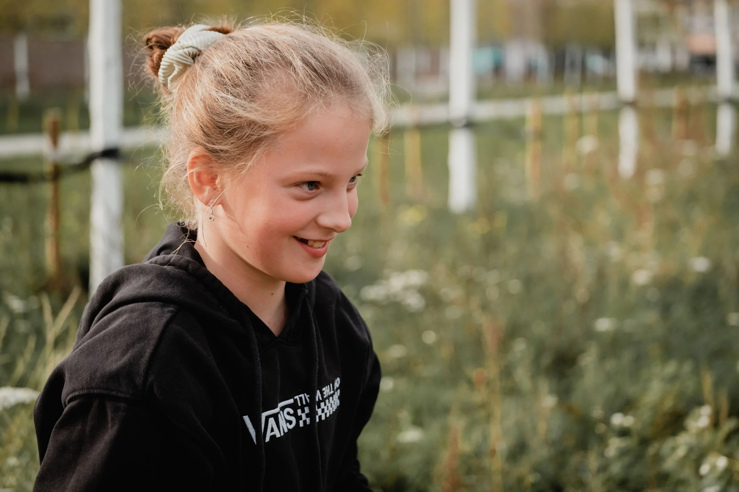 A young woman with light brown hair tied in a bun, smiling, wearing a black hoodie, outdoors near a body of water with trees in the background.