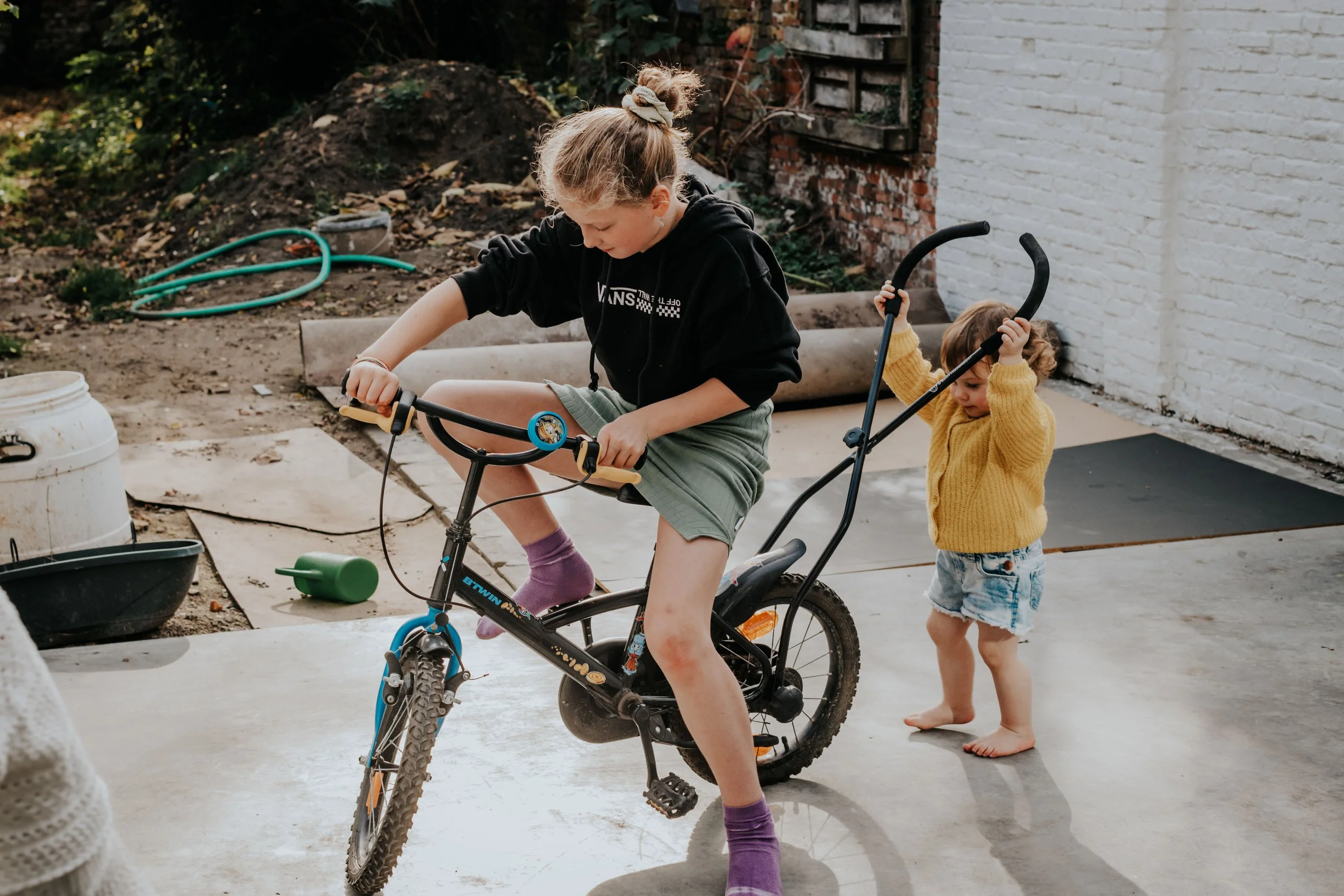A young girl riding a bicycle with training wheels and a curly-haired toddler walking behind her in a backyard.