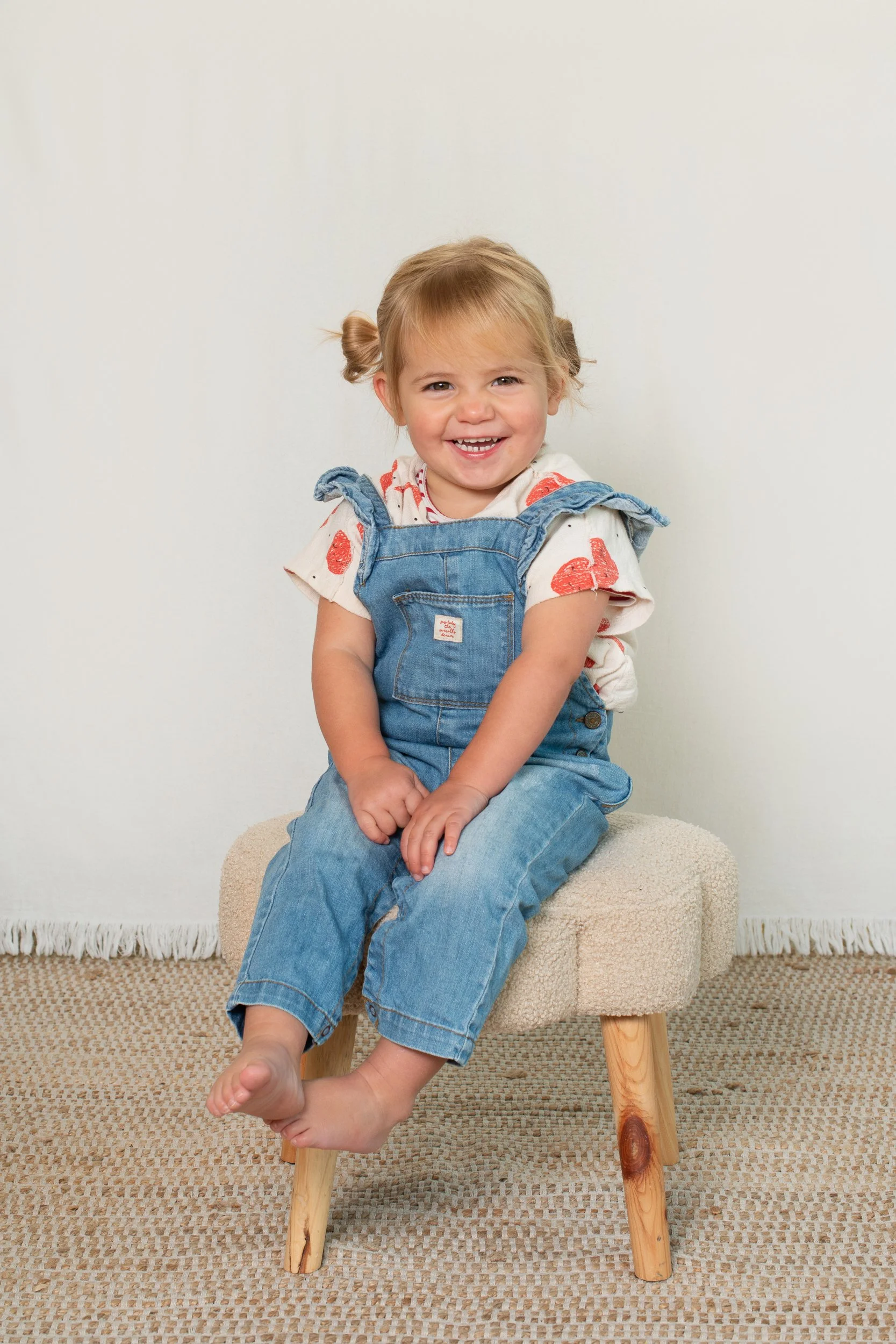 A smiling young girl with blonde hair tied in pigtails, wearing a white t-shirt with red patterns and denim overalls, sitting on a beige stool in a room with a white wall, a beige woven rug, and wooden legs visible on the stool.