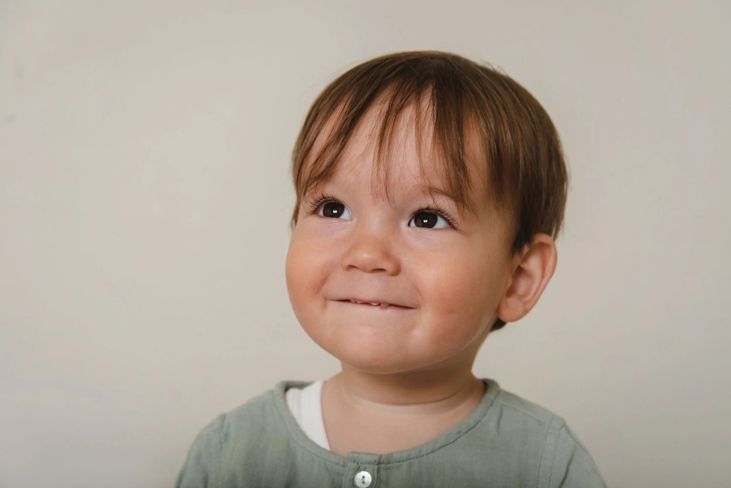 Close-up of a young boy with brown hair, dark eyes, smiling slightly, against a plain light-colored background, wearing a gray shirt with a white undershirt.