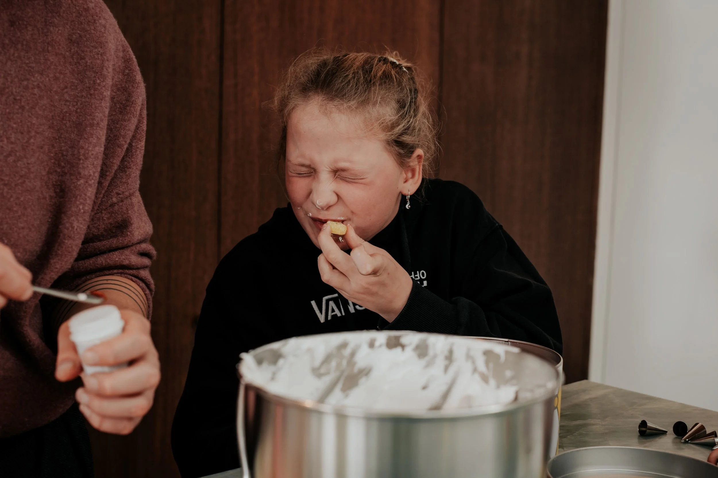 A young girl with curly hair and earrings is eating cake from a mixing bowl, with another person partially visible beside her.