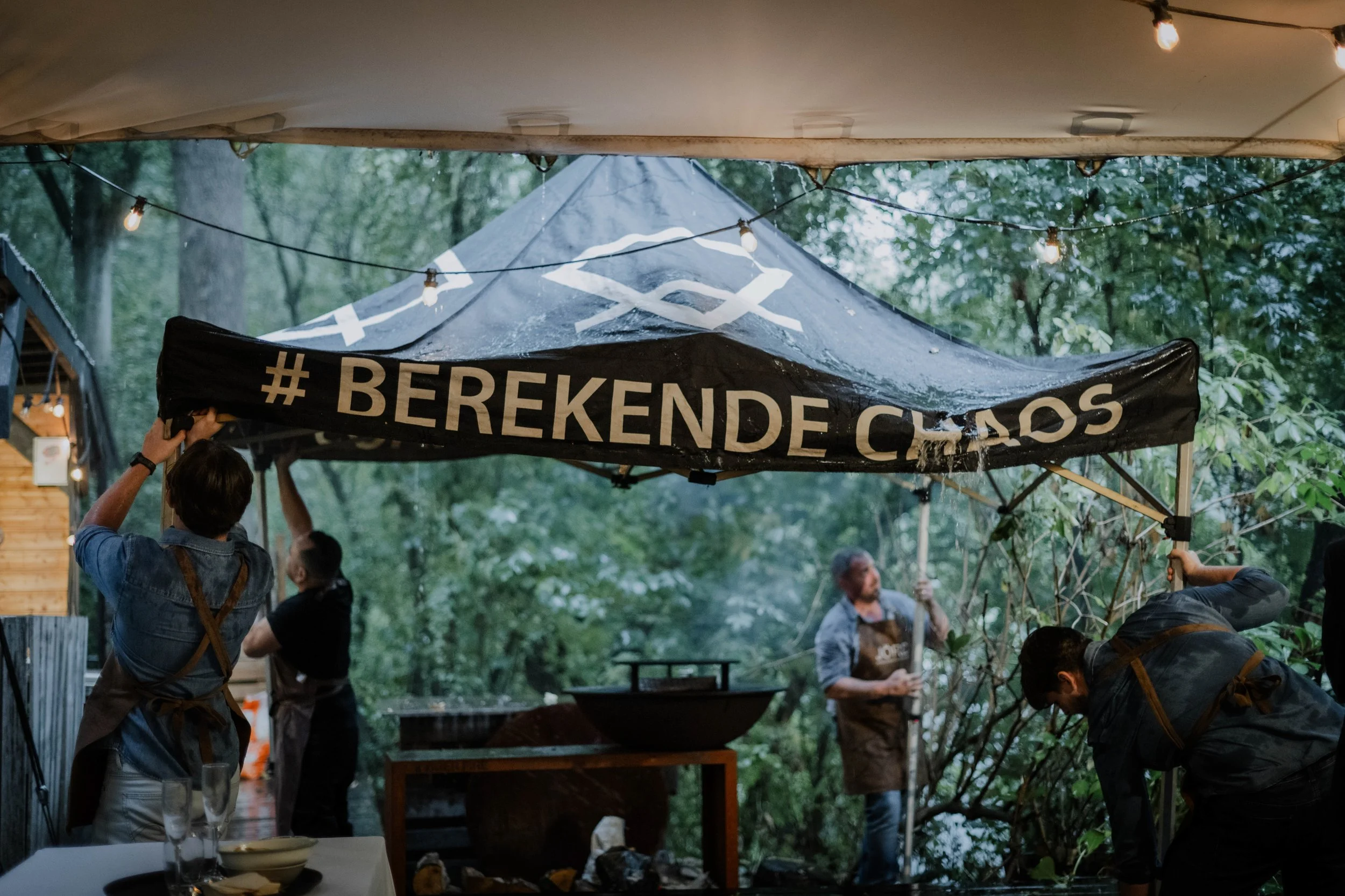 People setting up an outdoor event under a canopy in a wooded area, with string lights hanging and a black banner that reads '# BEREKENDE CASS'.