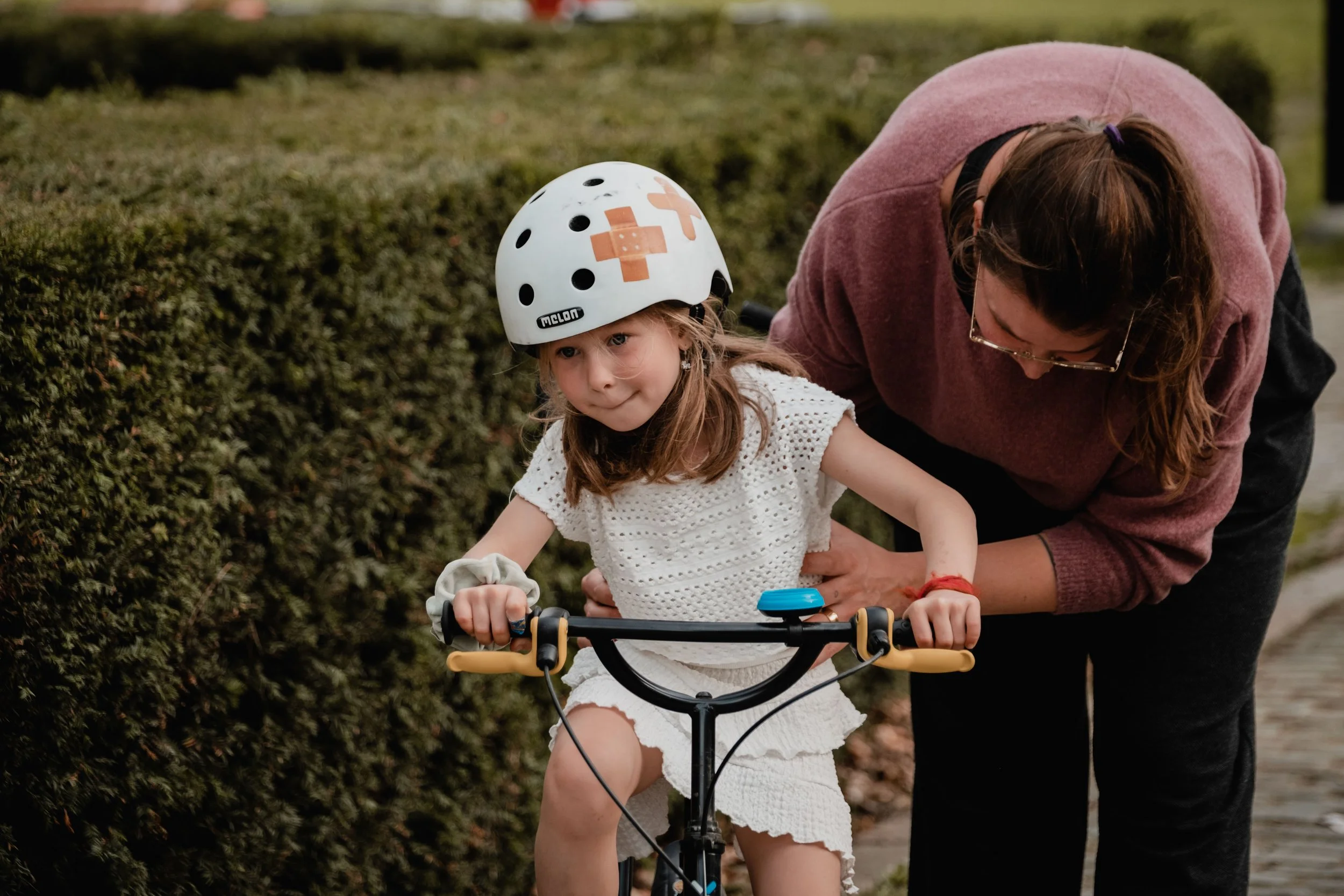 A young girl wearing a bicycle helmet and white dress riding a bike with help from a woman, likely her mother, on a trail beside a hedge.