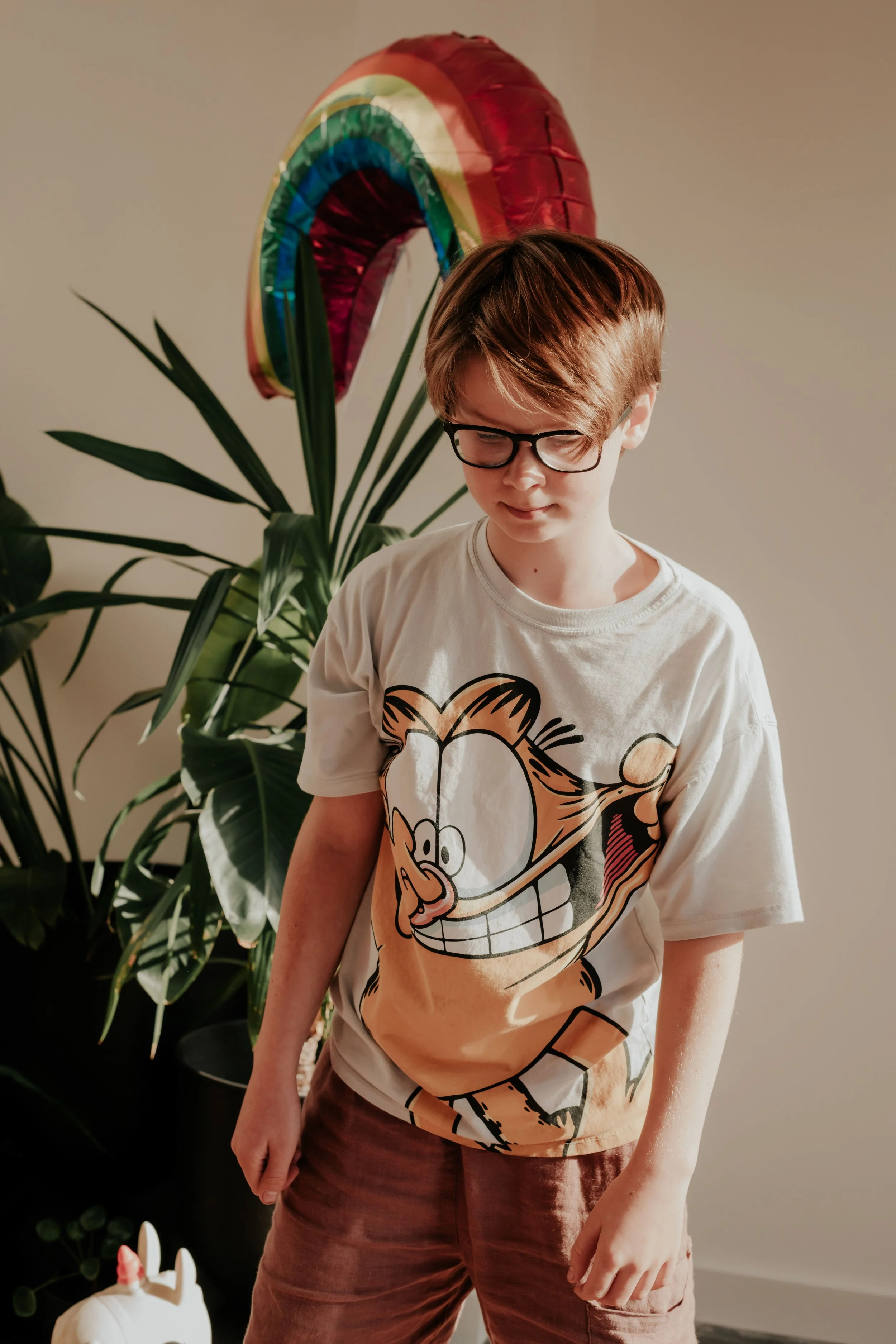 A young boy with brown hair and glasses standing in front of a wall with a large rainbow-shaped balloon. The boy is wearing a cartoon character T-shirt and brown pants.