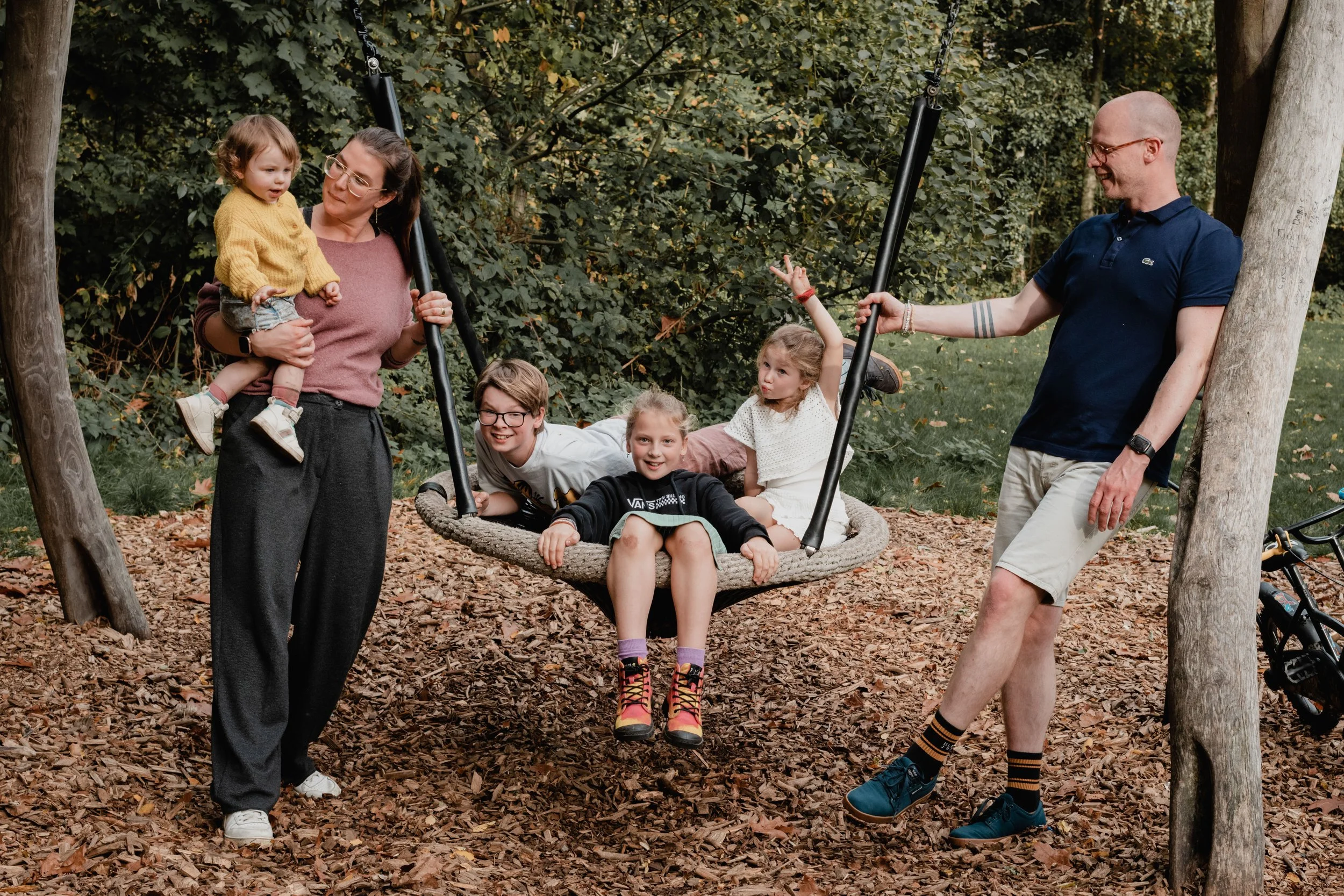 Family enjoying outdoor play on a swing set in a wooded area, with children on the swing and parents nearby.