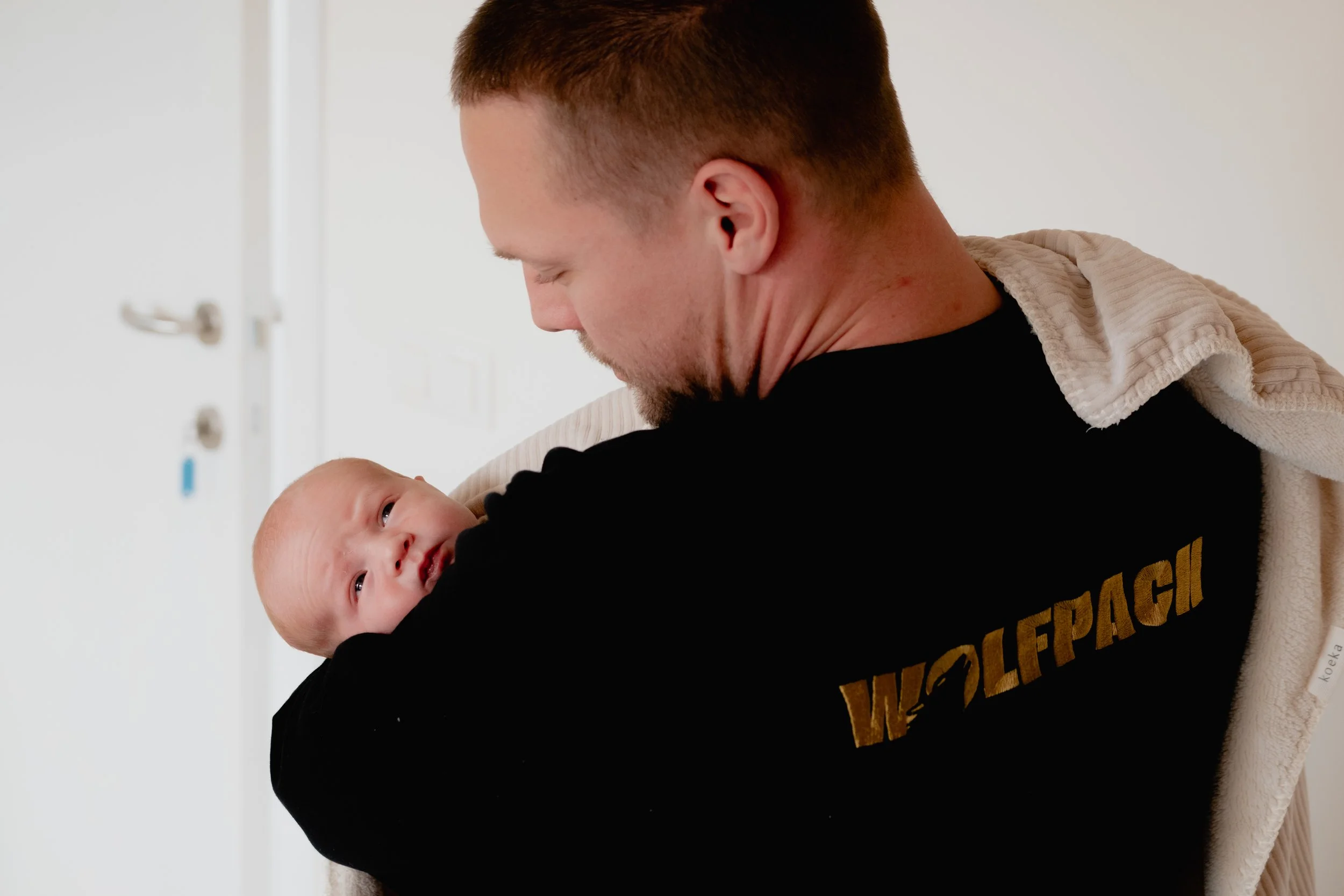 A man holding a newborn baby in a medical setting, with a white wall and medical equipment in the background.