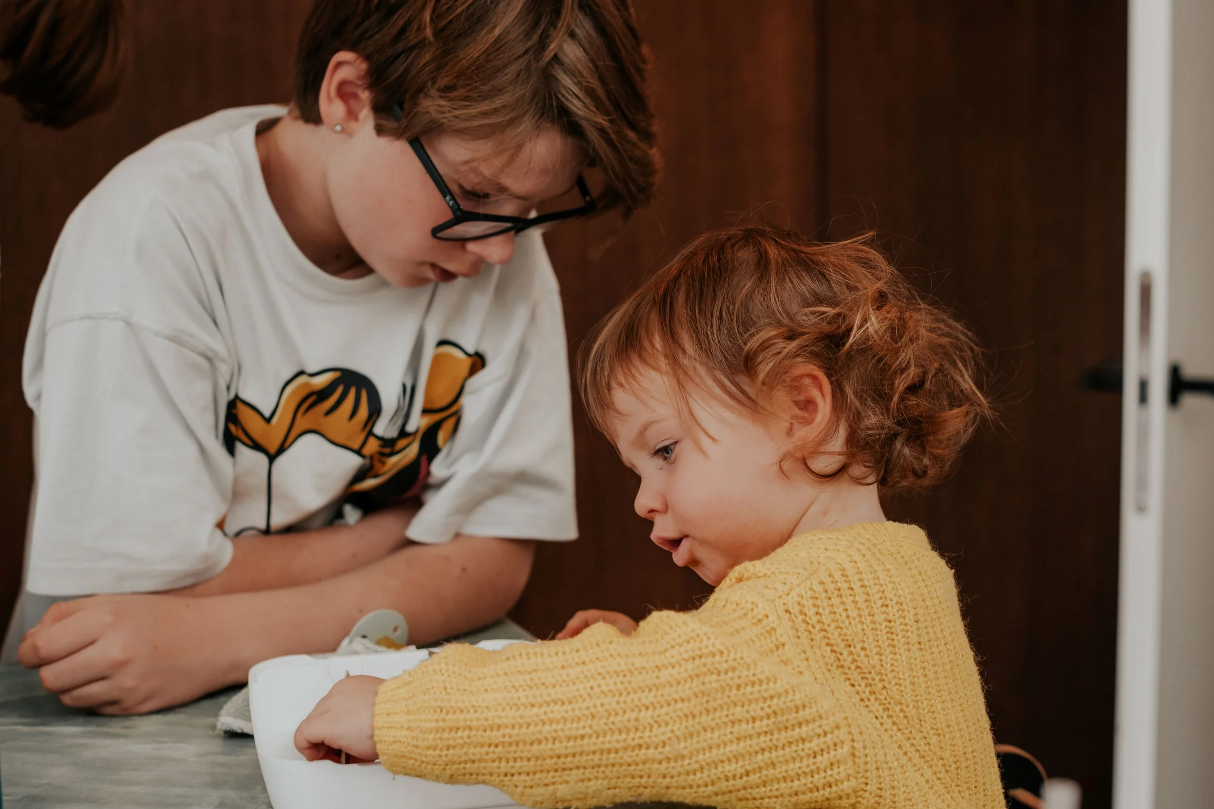 A woman with short brown hair and glasses, wearing a white T-shirt with a tiger design, leans over and assists a young girl with curly red hair and a yellow sweater, both focused on a table with a white device.