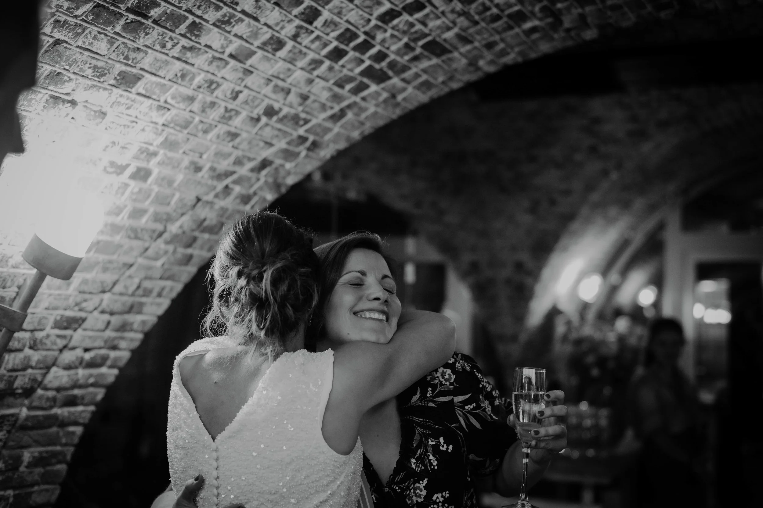 Two women, one wearing a white dress and the other in a darker floral dress, hugging and smiling at each other in a cozy, brick-ceiling restaurant or bar. One woman holds a glass of champagne.