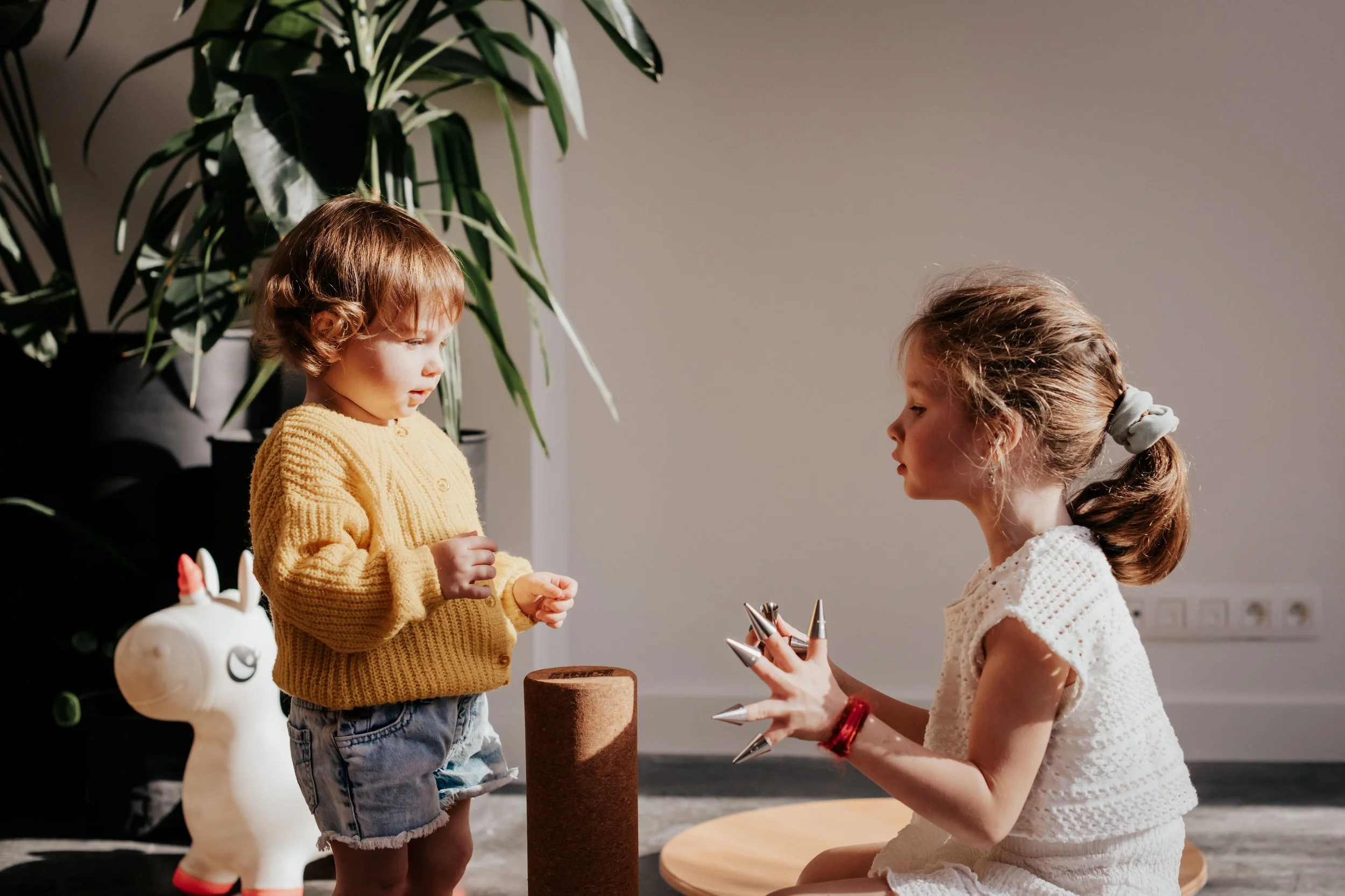 Two young girls with light brown and blonde hair interacting indoors. One girl is wearing a yellow sweater, and the other girl is dressed in a white, textured dress with a ponytail tied with a scrunchie. They are standing next to a round table with m