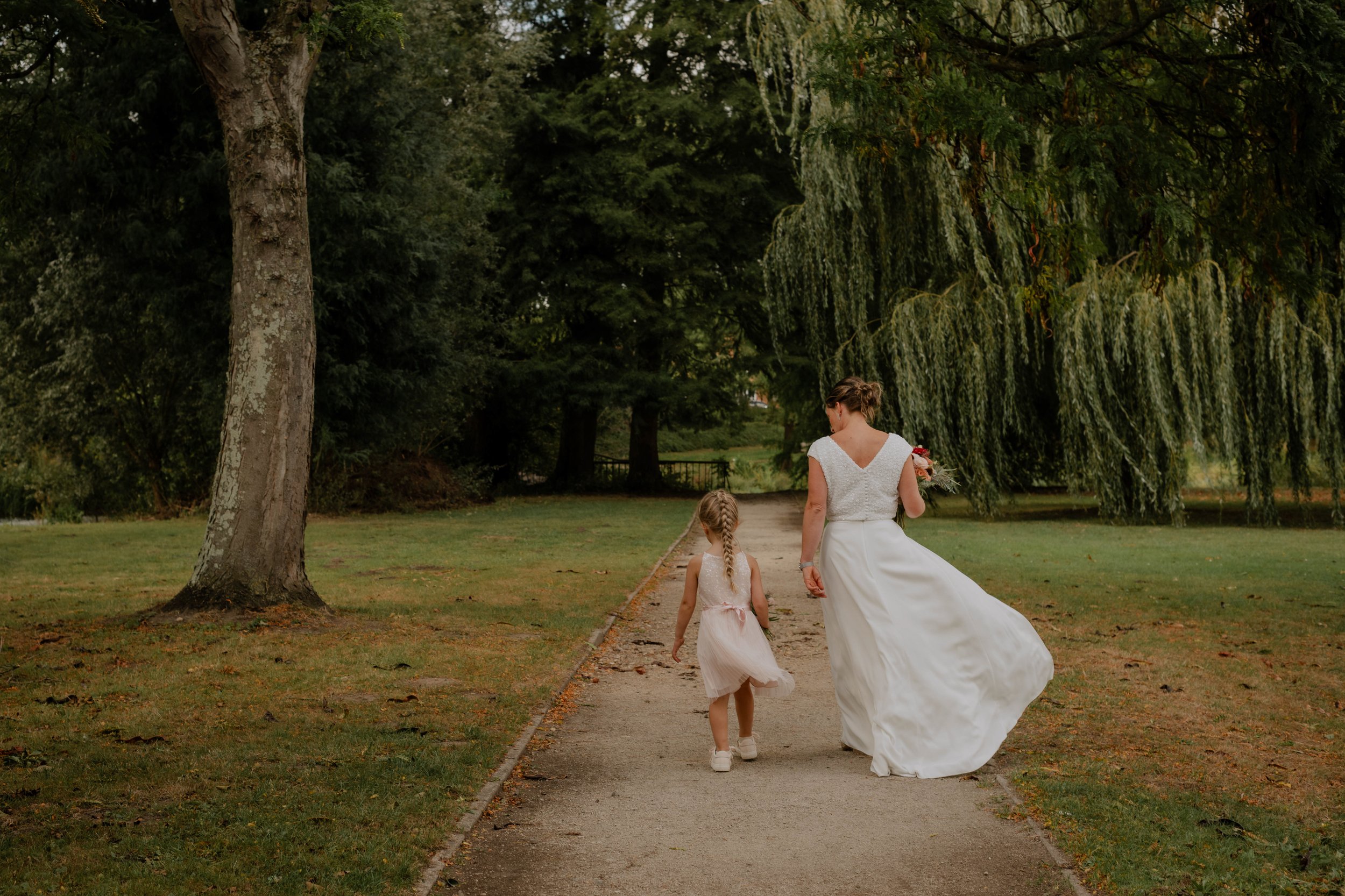 A woman in a white wedding dress walking hand in hand with two young girls, possibly bridesmaids or flower girls, on a park path surrounded by lush green trees.