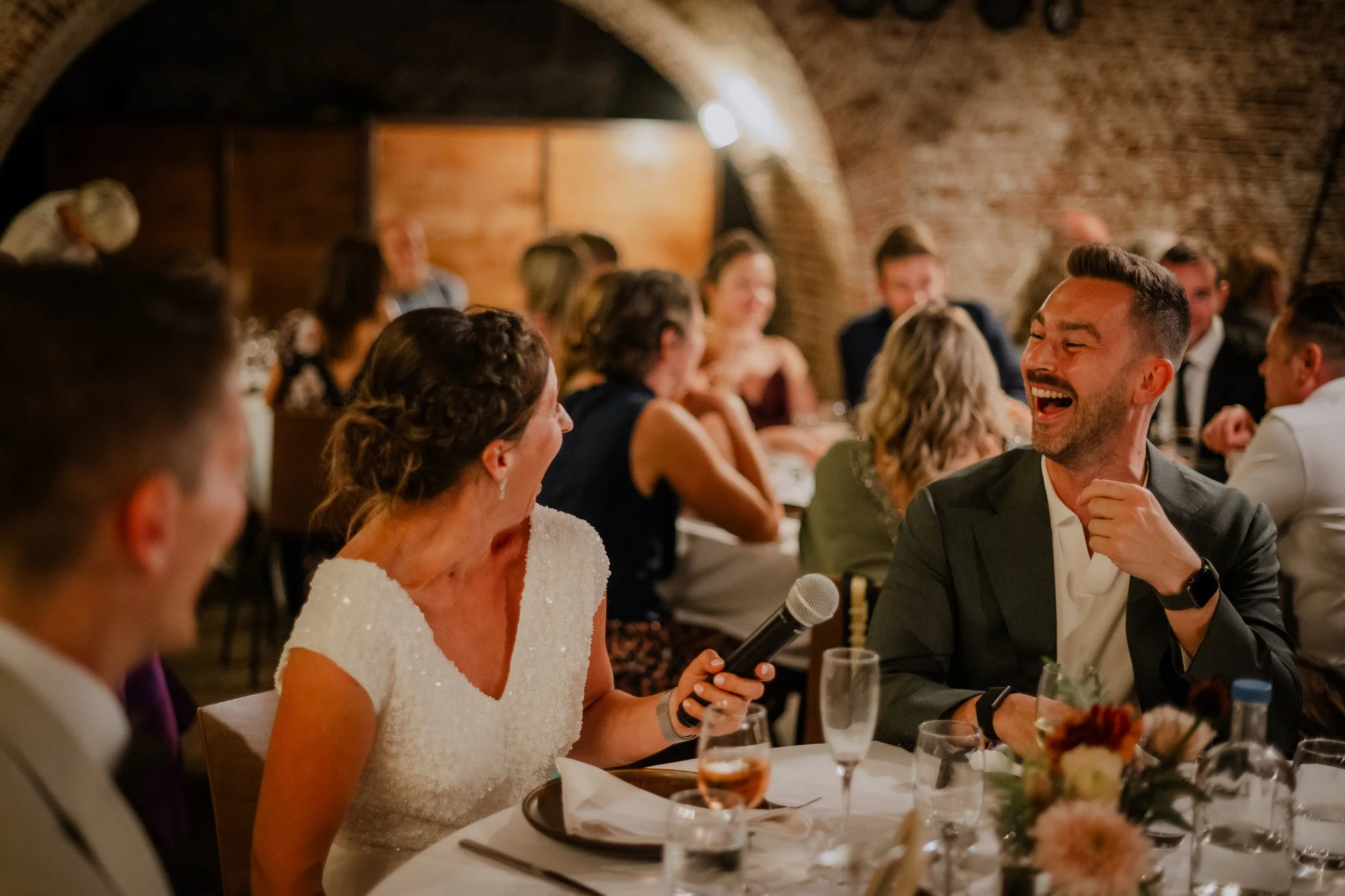 A woman in a white dress holding a microphone is speaking at a formal event. She is surrounded by people seated at a banquet table, with others smiling and laughing in the background. The setting is an elegant venue with brick arches.