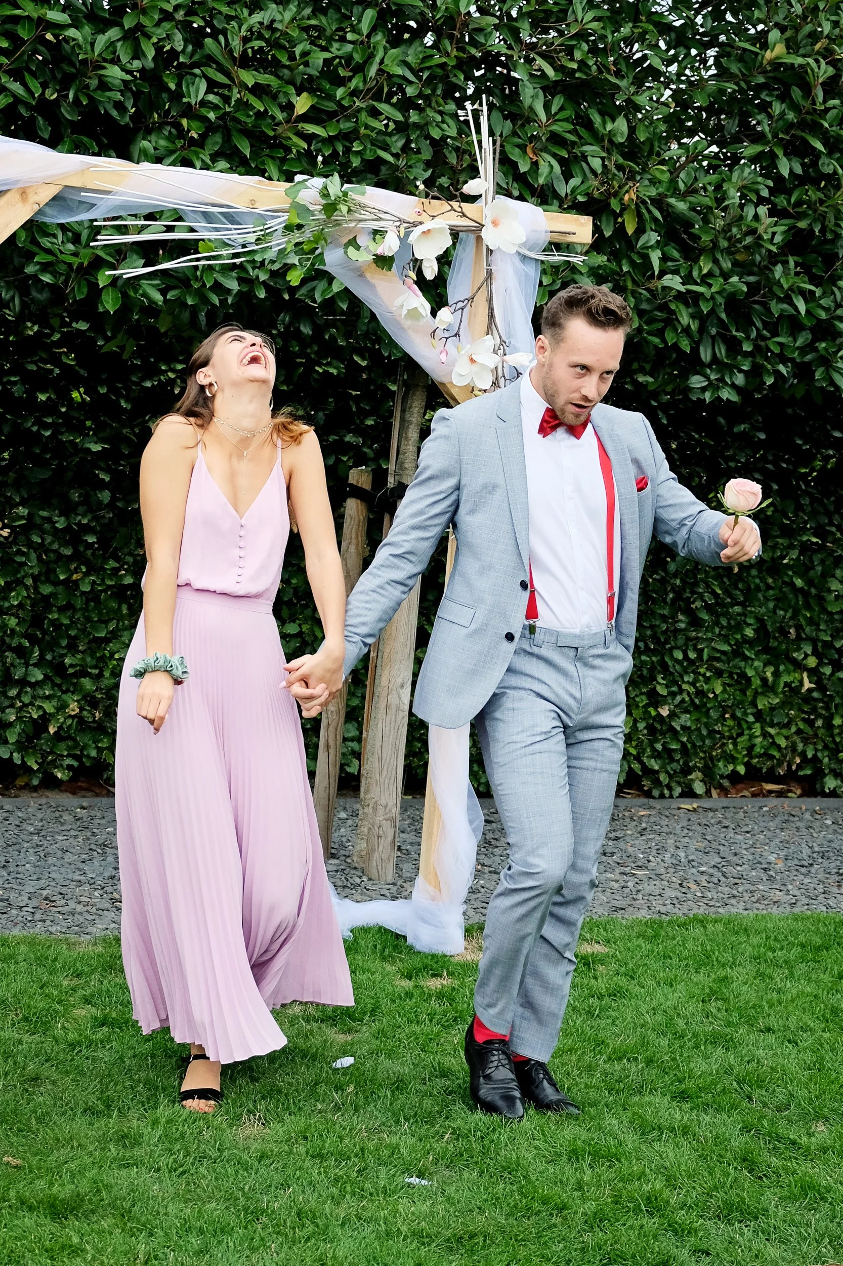 A couple holding hands at an outdoor wedding or celebration, standing in front of a decorative arch with flowers and tulle, surrounded by greenery.