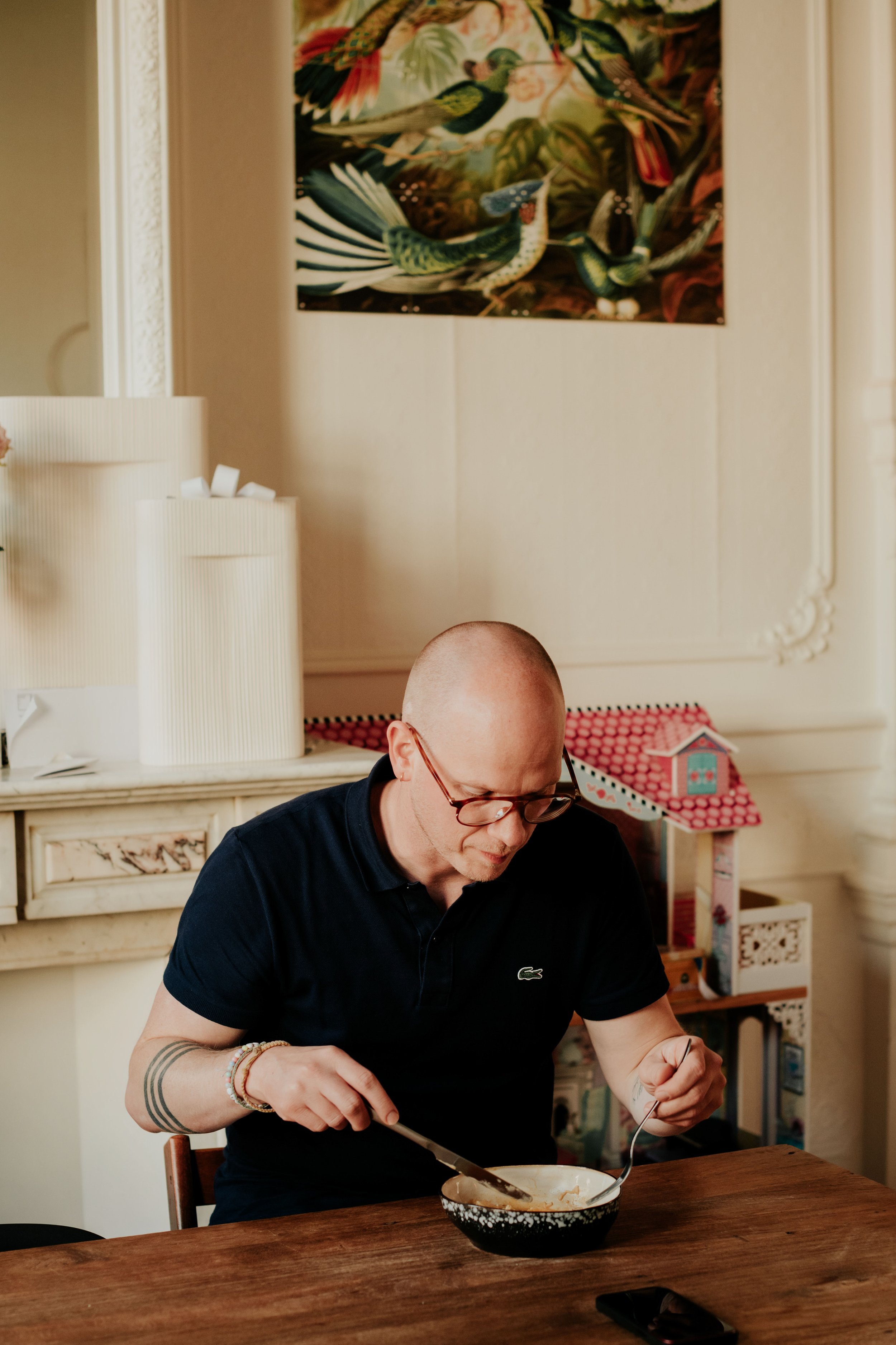 A bald man with glasses wearing a black Lacoste polo shirt sitting at a wooden table, eating food from a bowl with a spoon. There is a white cabinet with a lamp and a framed picture on the wall behind him.