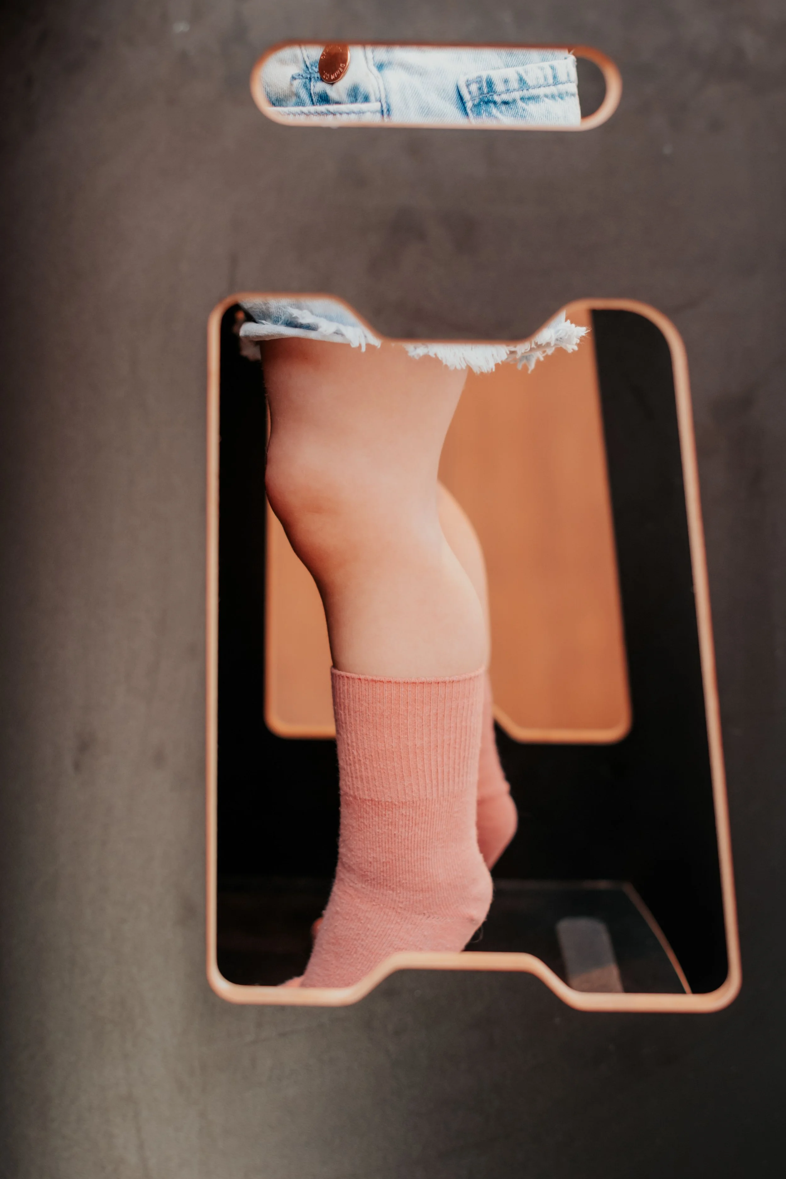 Close-up of a mirror reflecting a person's leg wearing a pink sock, with denim shorts and a copper button visible at the top of the reflection.
