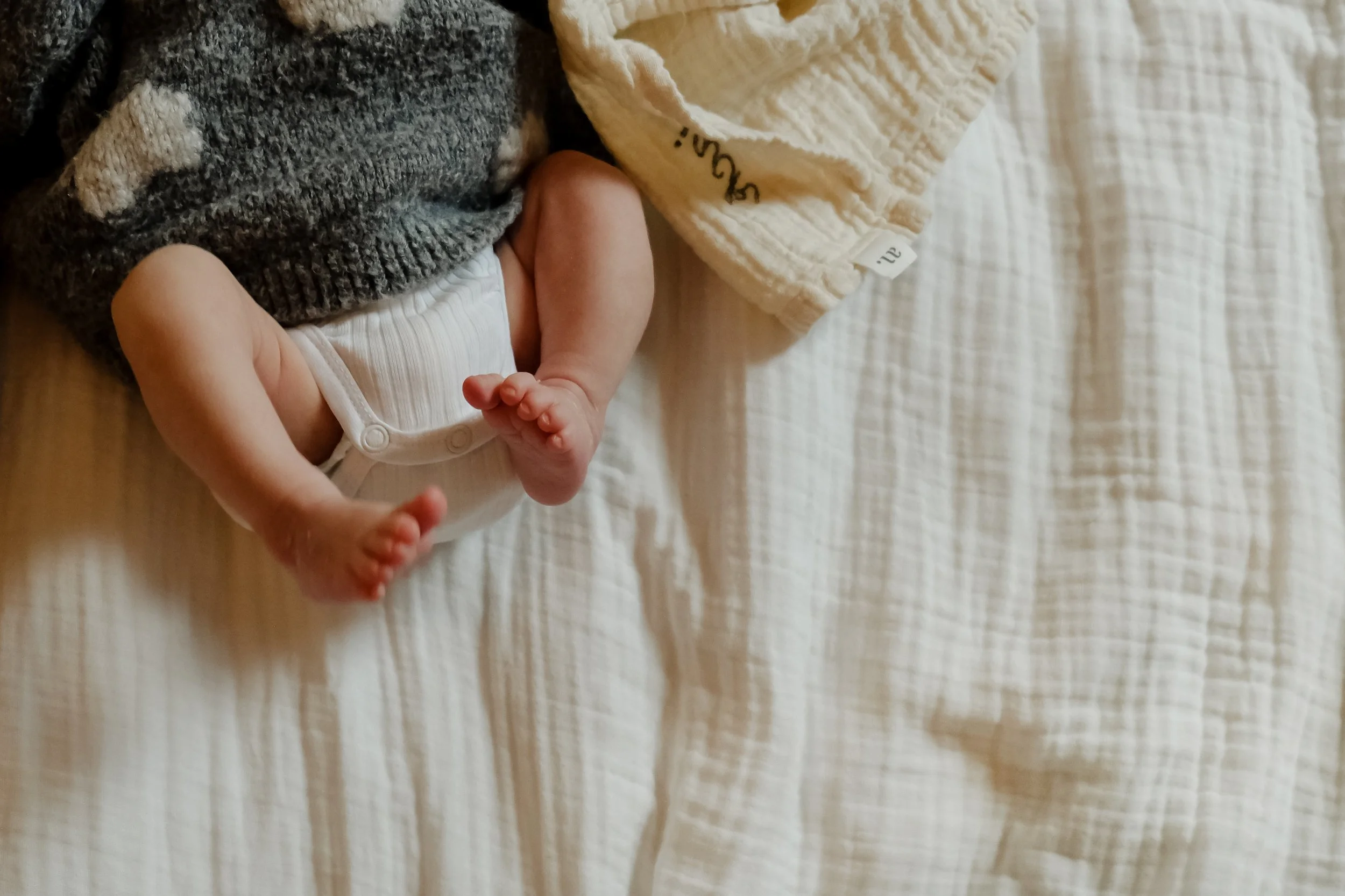Close-up of a baby's feet, with one foot partially covered by a gray knit sock and the other exposed, lying on a white textured blanket.