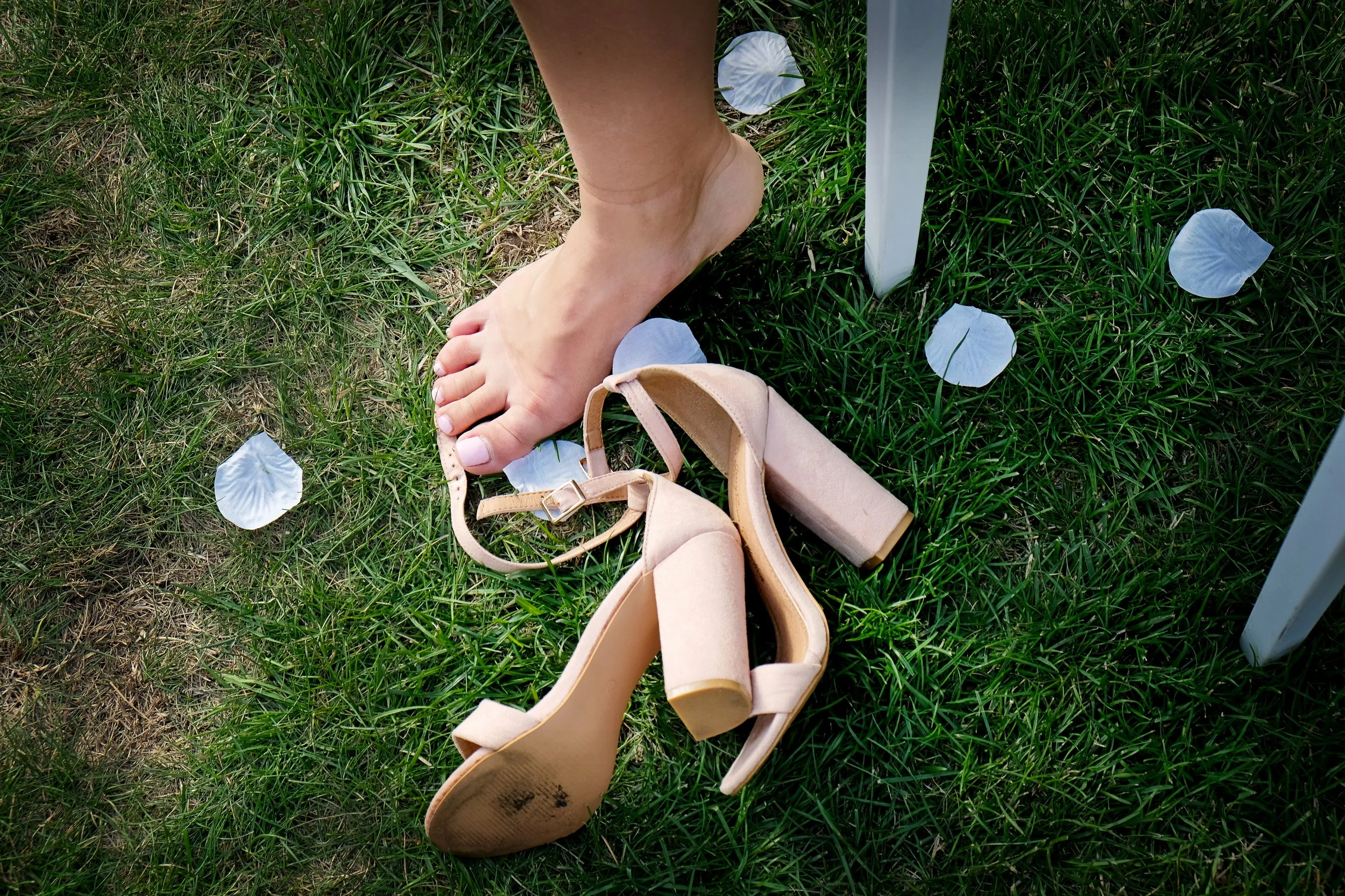 A person's bare foot on green grass surrounded by white flower petals and nude-colored high-heeled shoes, with a beige purse in the foreground.